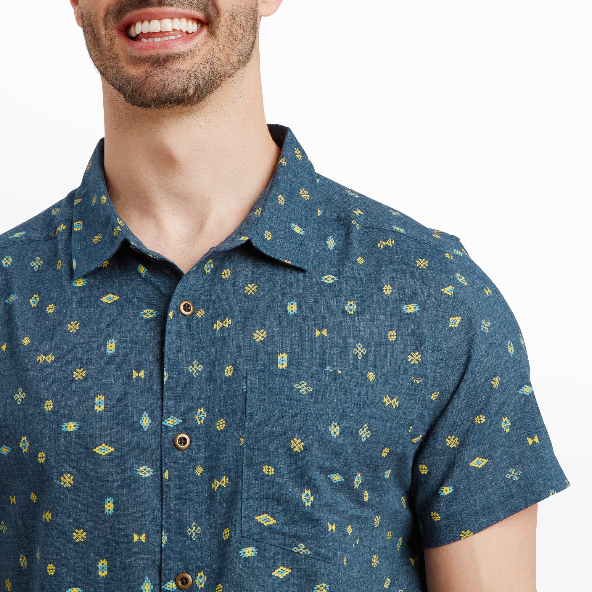 A close-up image of the Sherpa Adventure Gear Bhutan Geo Short Sleeve Shirt in Blue focuses on the upper chest area, showcasing the intricate geometric print and the quality stitching of the single chest pocket. The buttons and collar details are clearly visible, emphasising the craftsmanship and design of the garment.