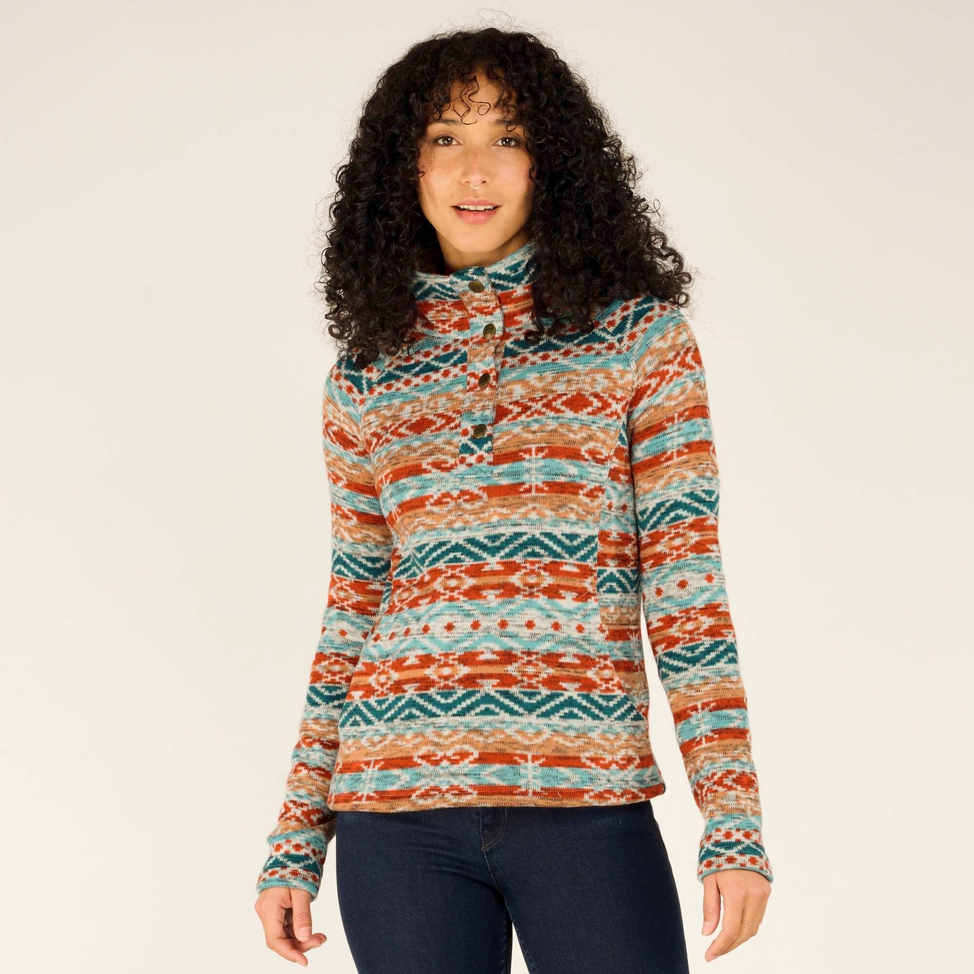 A woman with long, curly black hair wears Sherpa Adventure Gear Bhutan Pullover in Beige. The pullover has a high collar with buttoned detailing at the neckline. She stands facing the camera, slightly tilting her head with a relaxed expression. The pullover features intricate geometric patterns, and she pairs it with dark blue jeans. The background is neutral and softly lit.