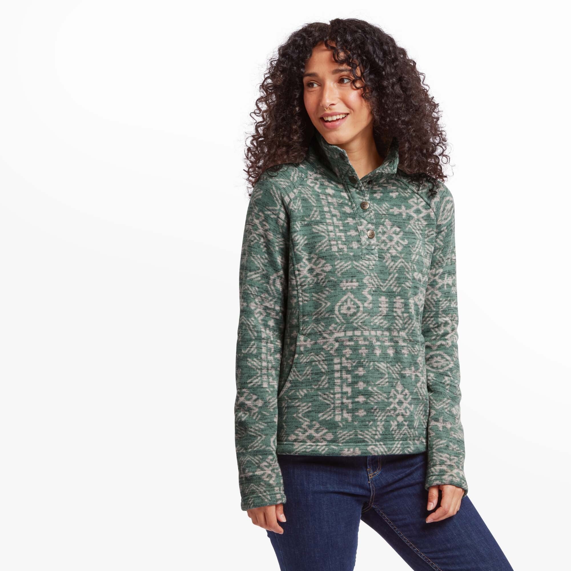 A young woman with curly dark hair and a warm smile is wearing a Sherpa Adventure Gear Bhutan Pullover in Green with a high buttoned collar. The sweater features an intricate white geometric pattern inspired by traditional Bhutanese designs. She stands slightly angled to the right with a relaxed posture, hands at her sides, wearing dark blue jeans.