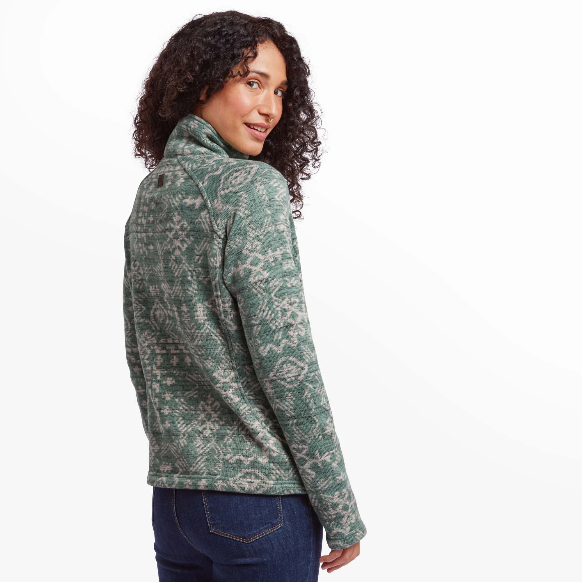 A back view of the model wearing the Sherpa Adventure Gear Bhutan Pullover in Green, slightly turning her head to the left to smile at the camera. The high collar remains upright, and the intricate geometric pattern continues across the back of the sweater. The fabric appears soft and textured.