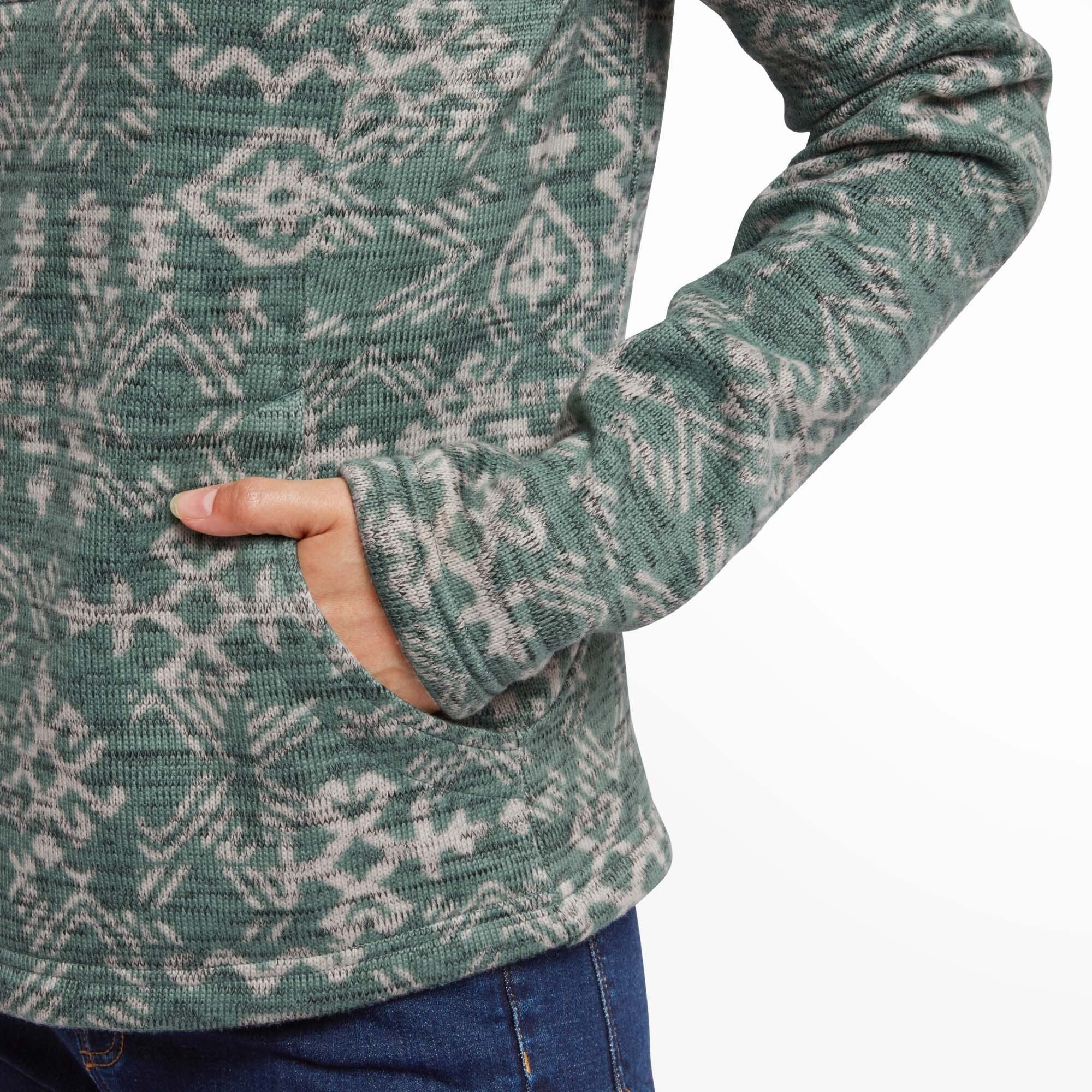 A close-up of the kangaroo pocket on the Sherpa Adventure Gear Bhutan Pullover in Green, with the model’s hand resting inside. The pattern aligns seamlessly across the front of the sweater, and the sleeve extends slightly over her wrist, emphasizing a cozy and comfortable fit.