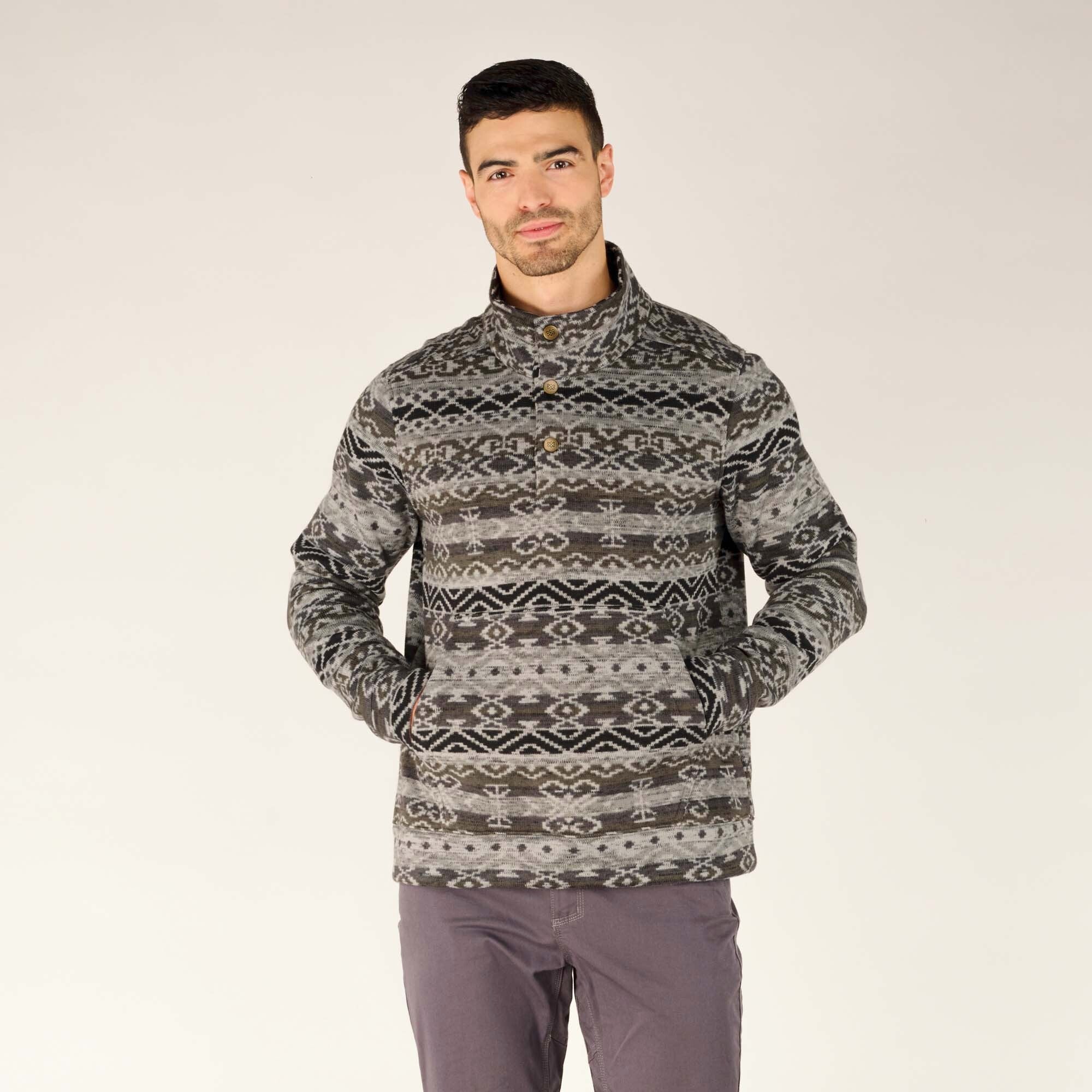 A man wearing a Sherpa Adventure Gear Bhutan Pullover in Grey pattern. The pullover features a stand-up collar with three buttons and a symmetrical geometric design throughout. The model has his hands in the pullover's kangaroo-style pocket, paired with dark grey trousers.