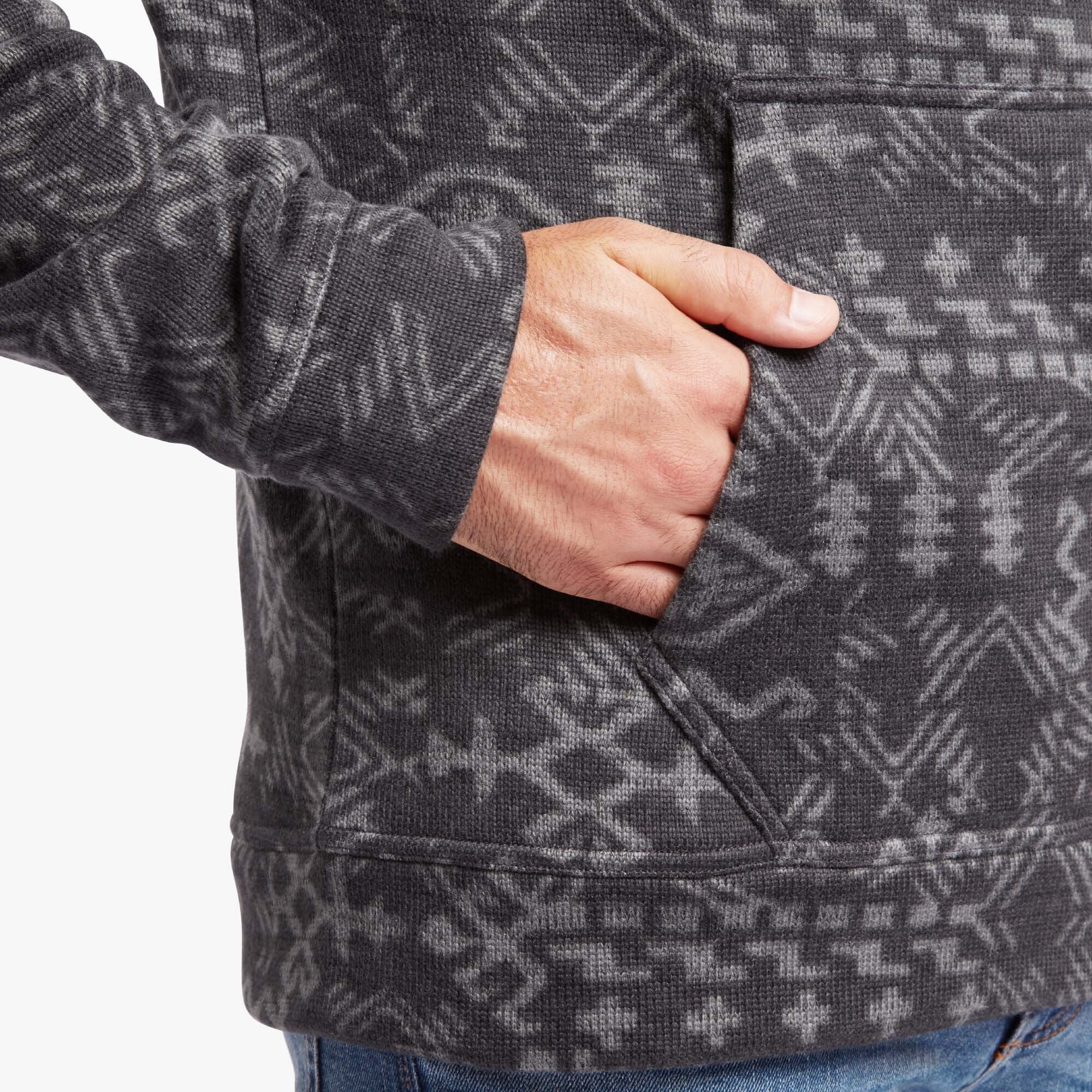 A close-up image of a person wearing a Sherpa Adventure Gear Bhutan Pullover in Grey with intricate light grey Bhutan-inspired geometric patterns. The focus is on the lower torso and one hand, which is casually placed inside the kangaroo pocket. The ribbed cuff of the sleeve is visible, highlighting the soft, textured fabric. The pullover is paired with blue jeans, showcasing a relaxed and cozy look. The detailed patterns add cultural and aesthetic interest to the garment.