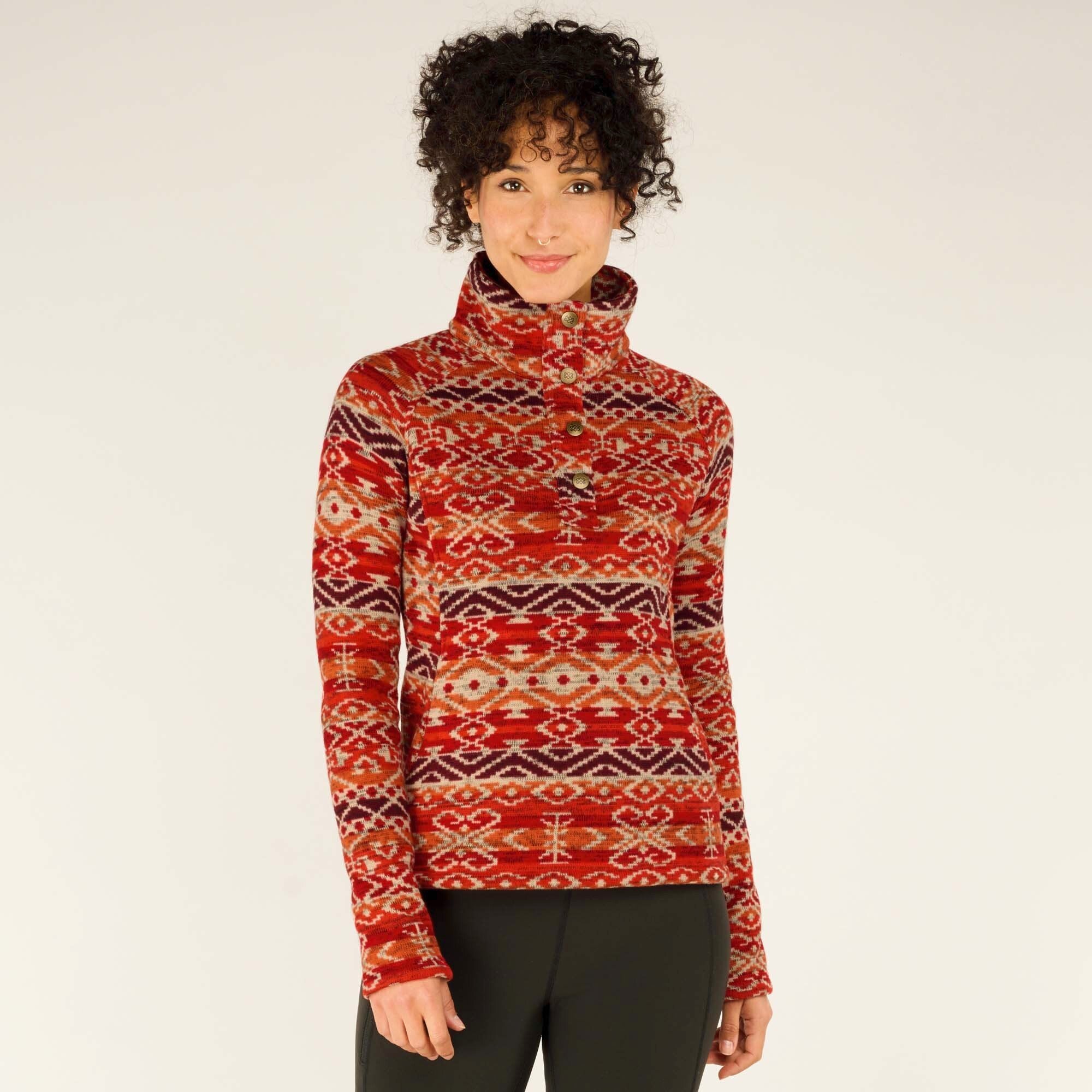 A woman with curly black hair models a Sherpa Adventure Gear Bhutan Pullover in Red with a high collar and buttoned placket. She stands facing forward with a gentle smile, her arms relaxed at her sides. The pullover features intricate geometric designs in shades of red, burgundy, and beige. She wears black leggings, and the background is a simple off-white.