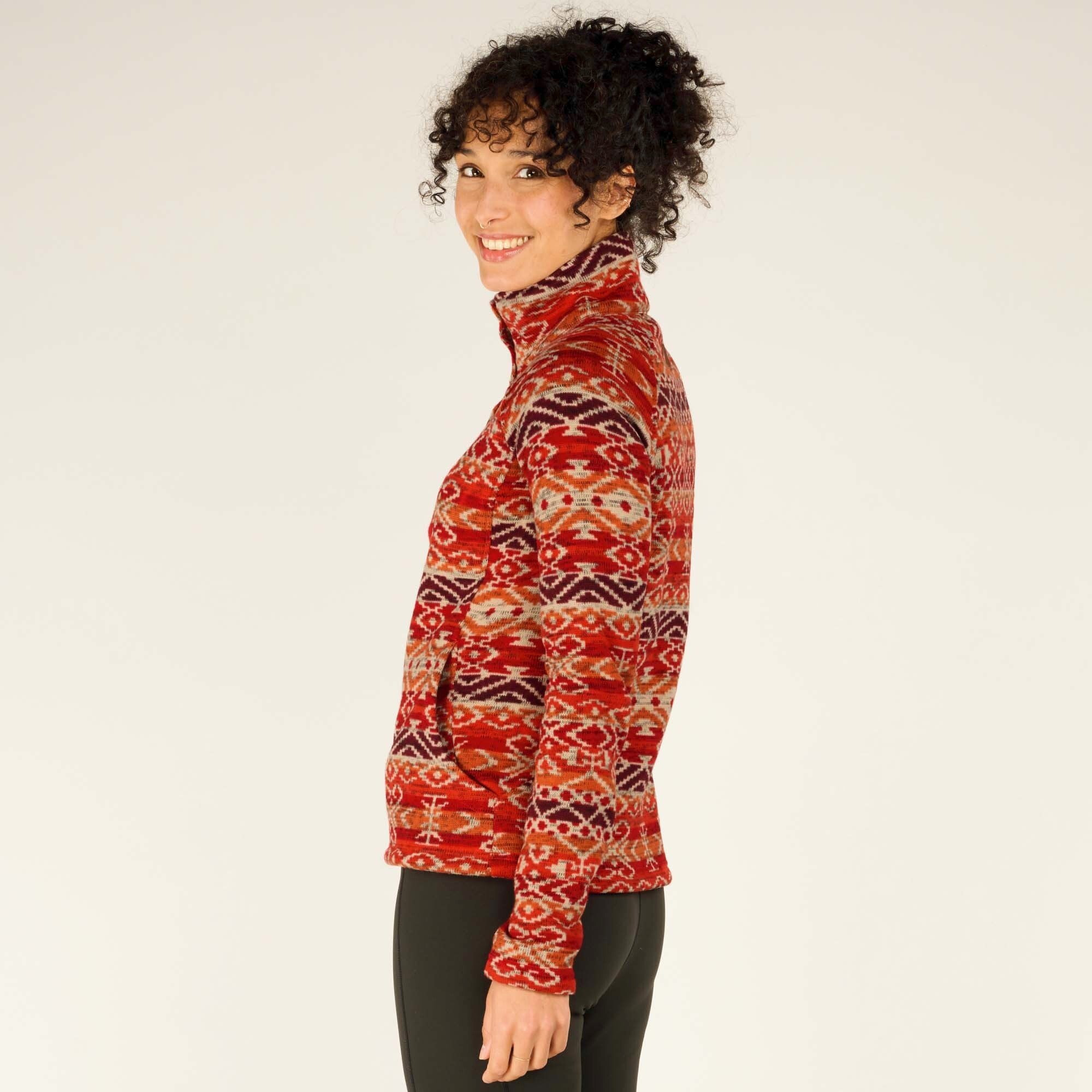 A side-profile view of the model wearing the Sherpa Adventure Gear Bhutan Pullover in Red, highlighting the high collar and the garment's well-fitted design. She is smiling brightly while looking slightly away from the camera. The intricate patterns in warm tones continue seamlessly on the sides of the pullover.