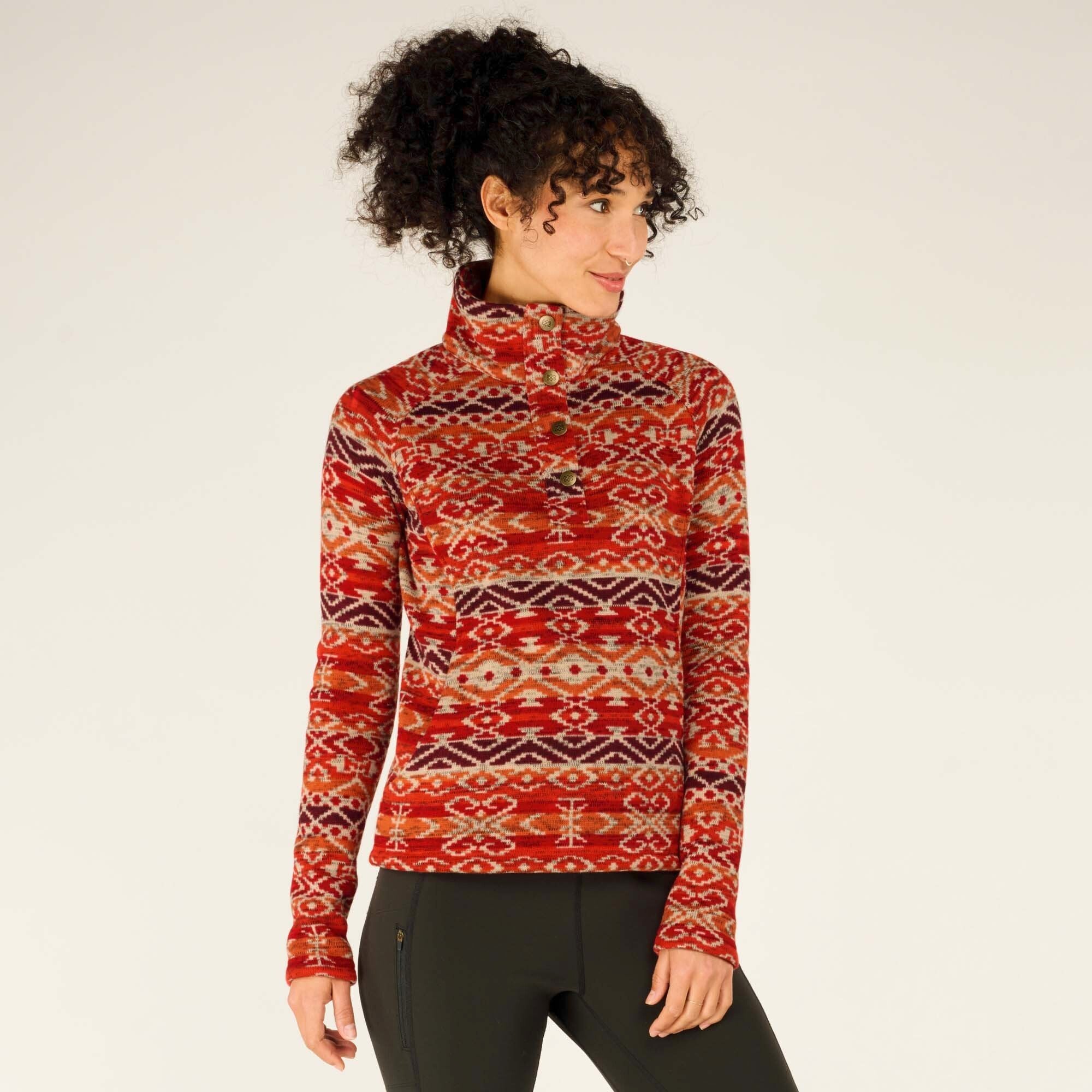 The model poses with her left hand slightly raised while turning her head to the left, offering a three-quarter view of the Sherpa Adventure Gear Bhutan Pullover in Red. She has a soft, content expression. The pullover's buttoned placket and high collar are prominent, and the fabric appears thick and warm.