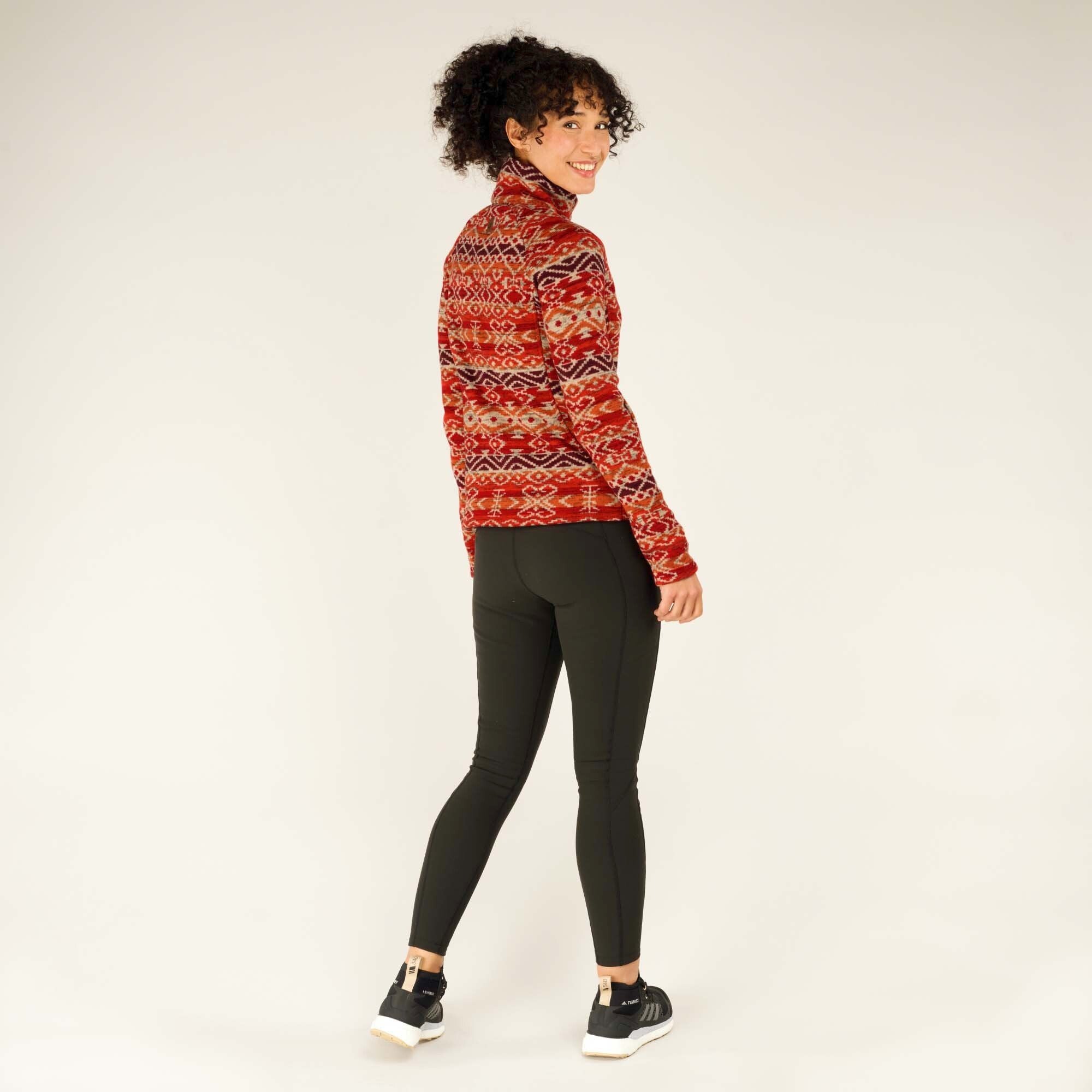 A dynamic full-body pose of the model, slightly turned away from the camera but looking back over her shoulder with a bright smile. Her black leggings and athletic shoes complement the Sherpa Adventure Gear Bhutan Pullover in Red, and her curly hair is slightly tousled. The pullover maintains a structured yet comfortable fit.