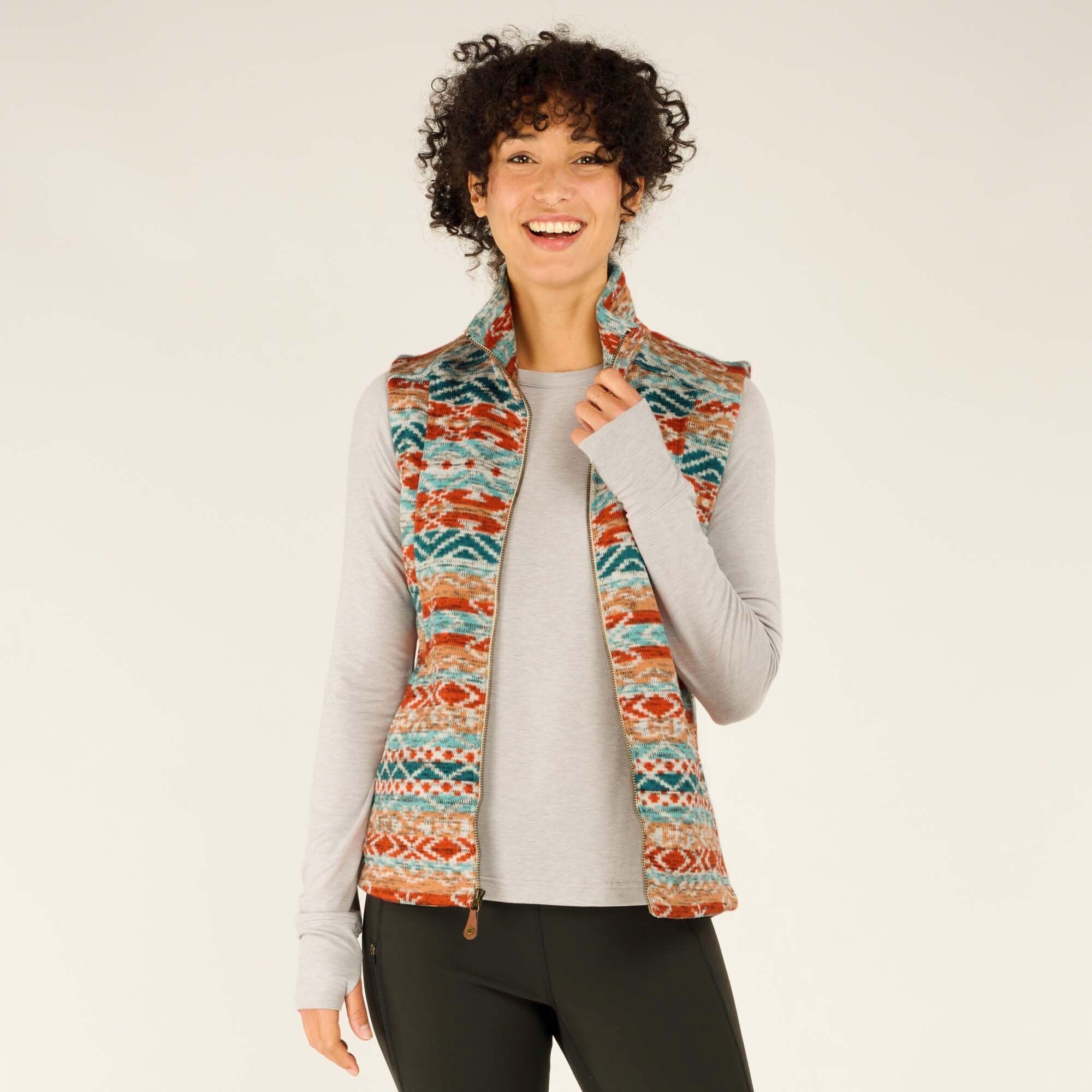 A woman with short curly hair and a bright smile poses wearing a Sherpa Adventure Gear Bhutan Vest in Beige with a vibrant, multicolored geometric pattern in shades of red, orange, teal, and cream. She wears a light gray long-sleeve top underneath and black fitted pants. The vest has a high collar and a full-length zipper. She gently holds the collar with one hand while standing in a relaxed pose against a neutral beige background.