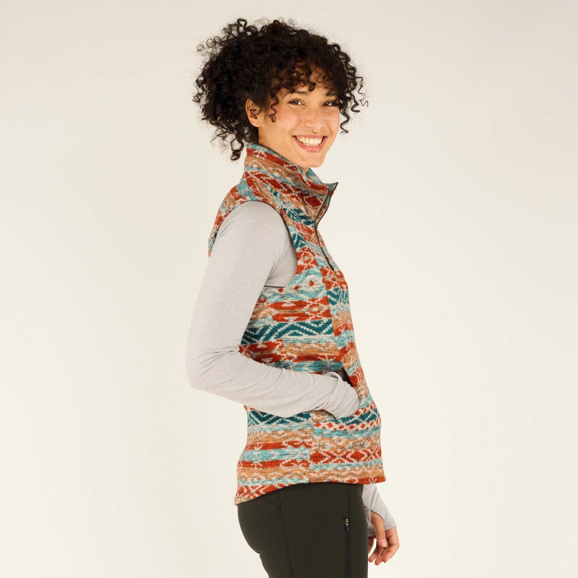 A close-up side profile of the woman wearing the Sherpa Adventure Gear Bhutan Vest in Beige. She smiles warmly at the camera, with her hands still resting in the vest’s pockets. The geometric pattern and texture of the fleece fabric are clearly visible. The high collar frames her face, and her curly hair is slightly tousled.