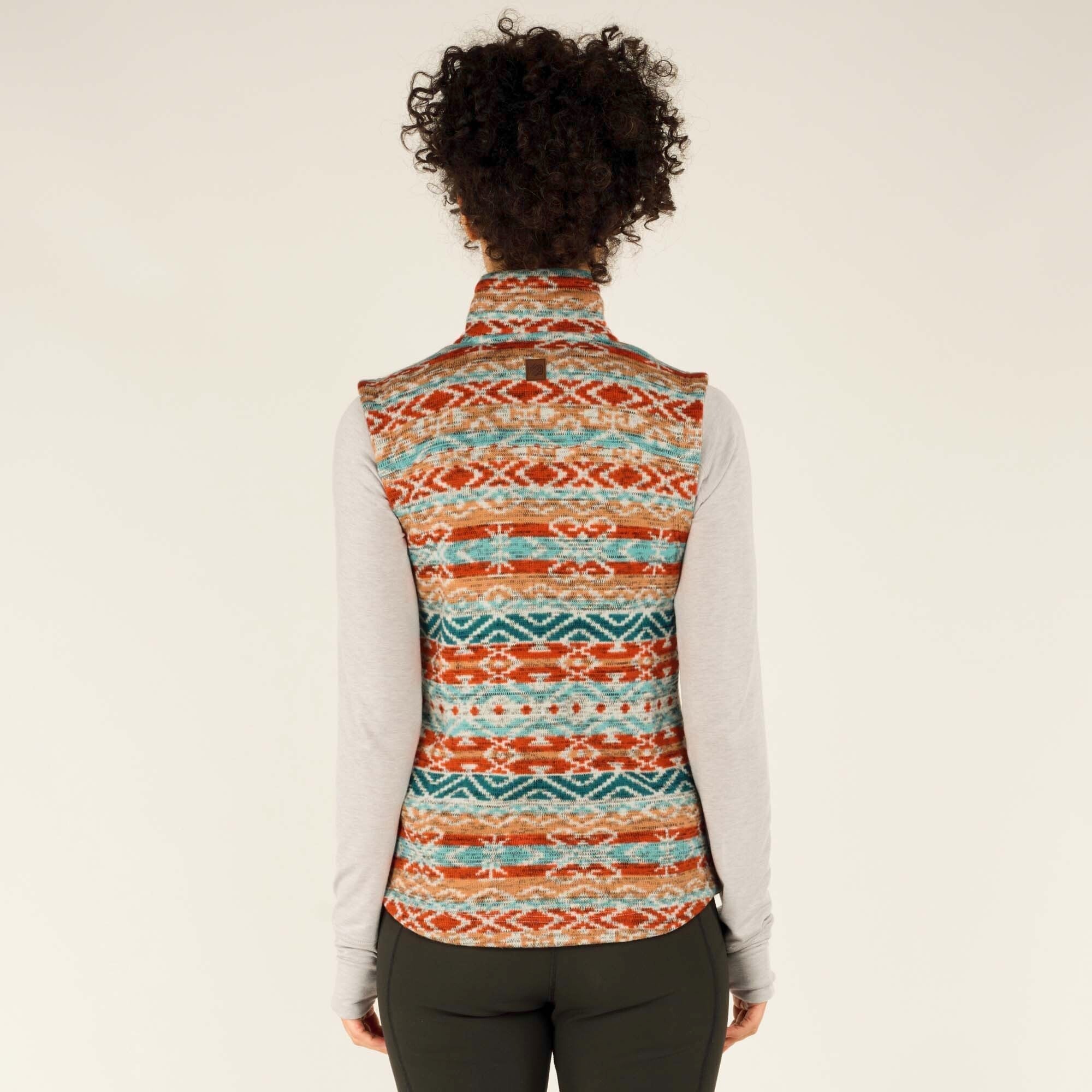 A rear view of the woman showcases the full back design of the Sherpa Adventure Gear Bhutan Vest in Beige. The multicolored geometric pattern continues seamlessly on the back. The vest has a slightly curved hem. She wears a long-sleeve gray top underneath, and her curly hair is slightly lifted, revealing the collar of the vest.