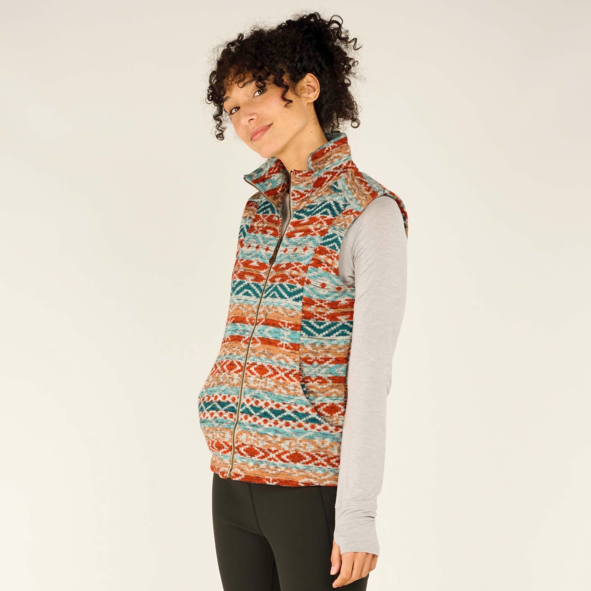 A three-quarter profile view of the woman, showing the front and side of the Sherpa Adventure Gear Bhutan Vest in Beige. She has a playful, relaxed expression with a slight smile. The vest’s texture and vibrant pattern stand out against her neutral-colored long-sleeve shirt and black leggings. The side pockets are slightly visible.