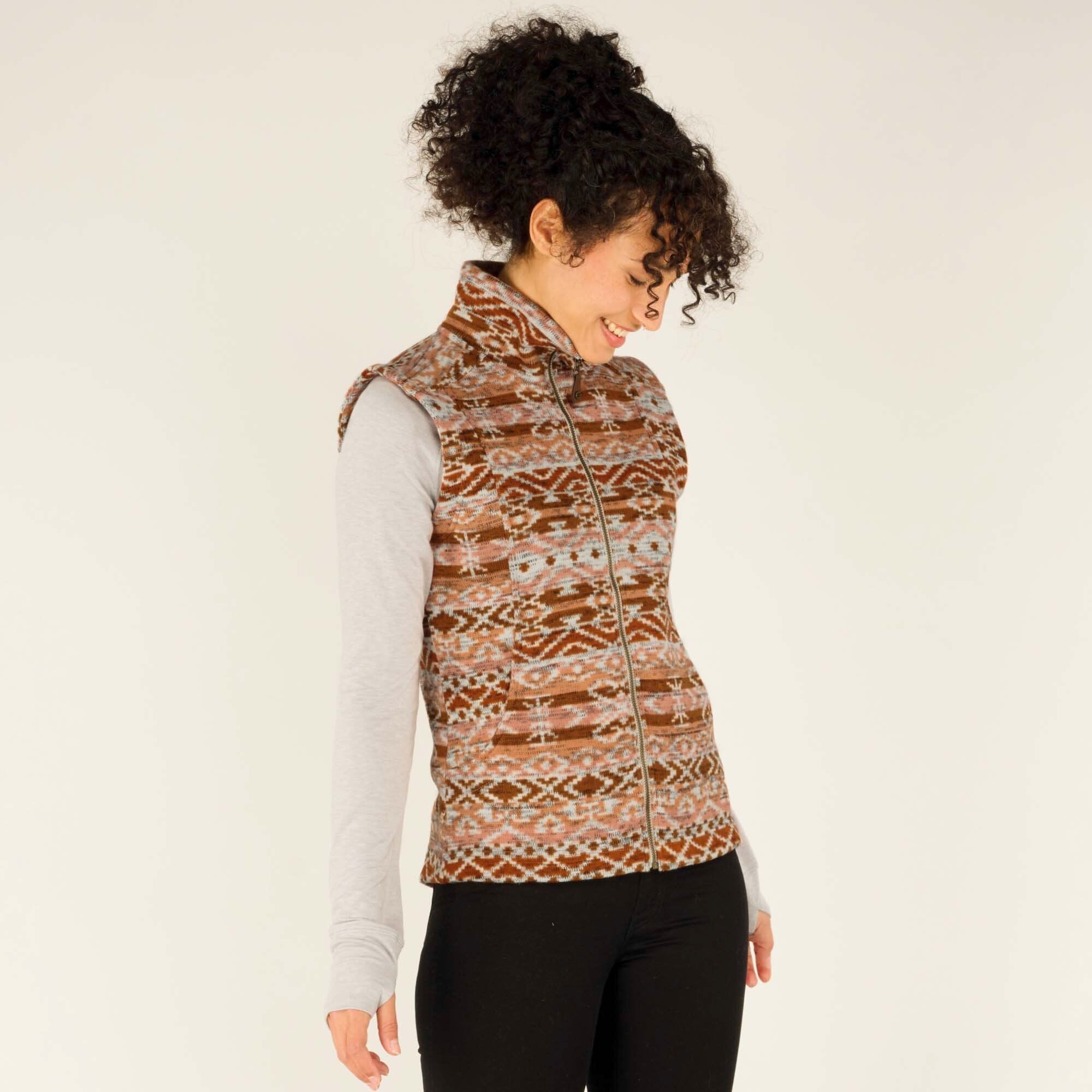 The model is turned slightly to her left, showcasing the side profile of the Sherpa Adventure Gear Bhutan Vest in Orange. She is smiling while looking down, with her curly hair pulled back. The vest’s high collar is visible, along with the detailed woven pattern. The long-sleeve gray shirt underneath contrasts with the vest, and her hands are relaxed by her sides.