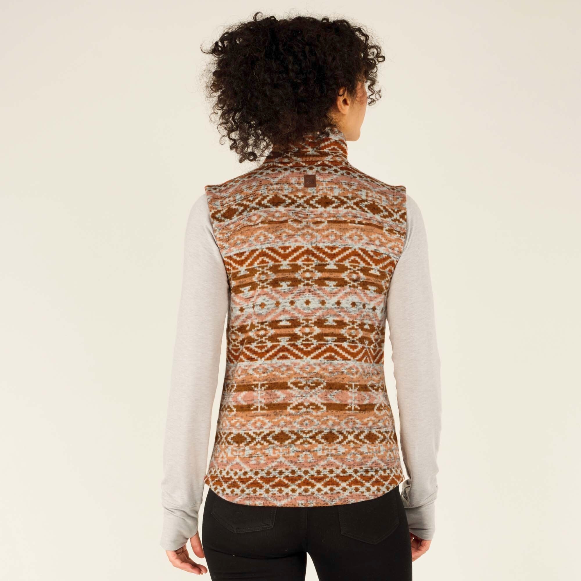 The model is facing away from the camera, displaying the full back of the Sherpa Adventure Gear Bhutan Vest in Orange. The Bhutanese-inspired geometric pattern continues seamlessly across the back, and the high collar stands up. The vest fits snugly, and the model's black leggings and long-sleeve gray shirt are also visible. Her curly hair is styled up, allowing for a clear view of the garment’s design.