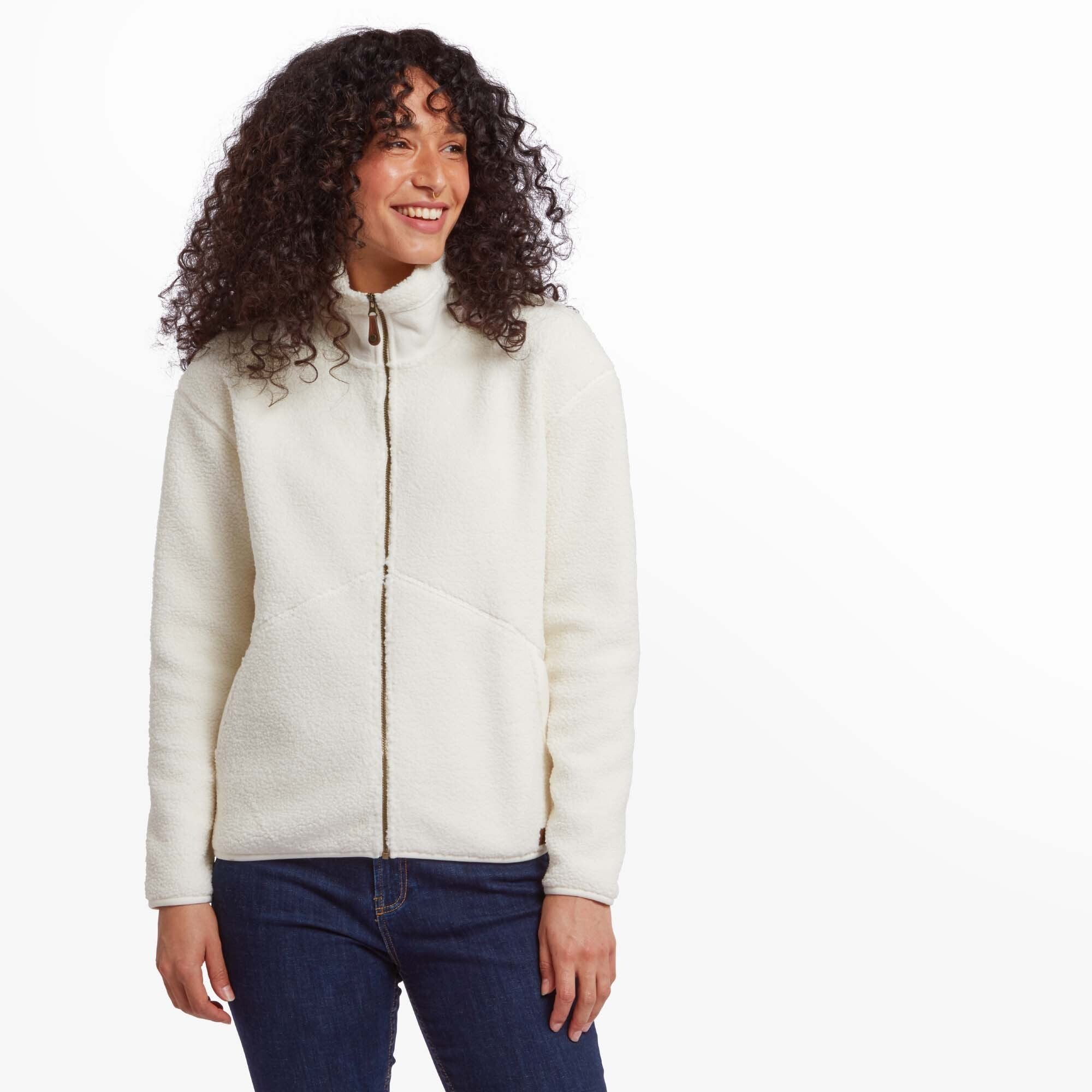 A woman wearing a Sherpa Adventure Gear Chamlang Full Zip Jacket in Cream with a full zip and a high collar. She is smiling warmly and pairing the jacket with dark blue jeans. The cozy texture of the fleece is prominently displayed, offering a casual and comfortable aesthetic.