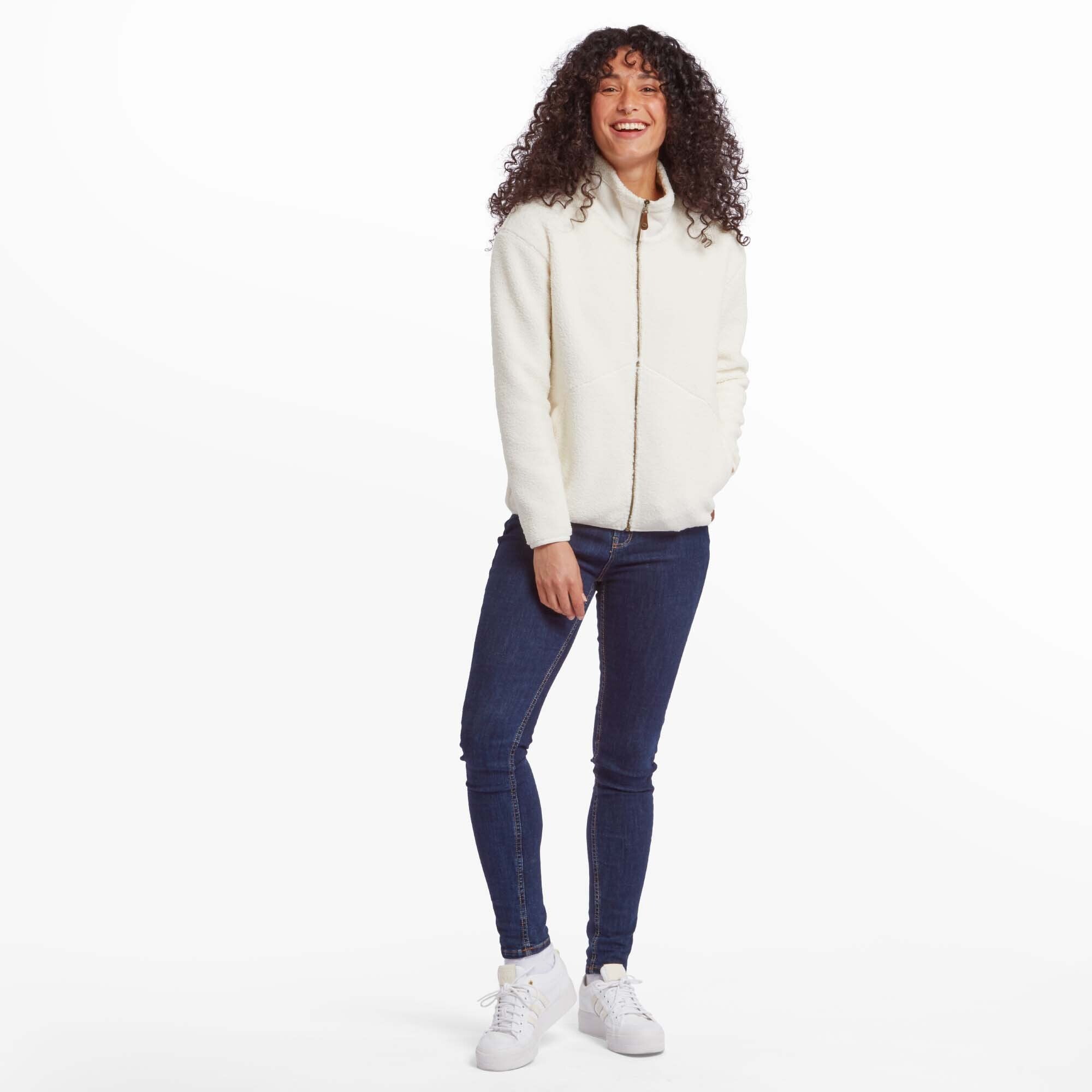 A full-body view of a woman wearing the Sherpa Adventure Gear Chamlang Full Zip Jacket in Cream paired with dark blue jeans and white trainers. She is standing casually with one hand in her pocket, smiling brightly, showcasing the jacket's relaxed fit and versatility for everyday wear.