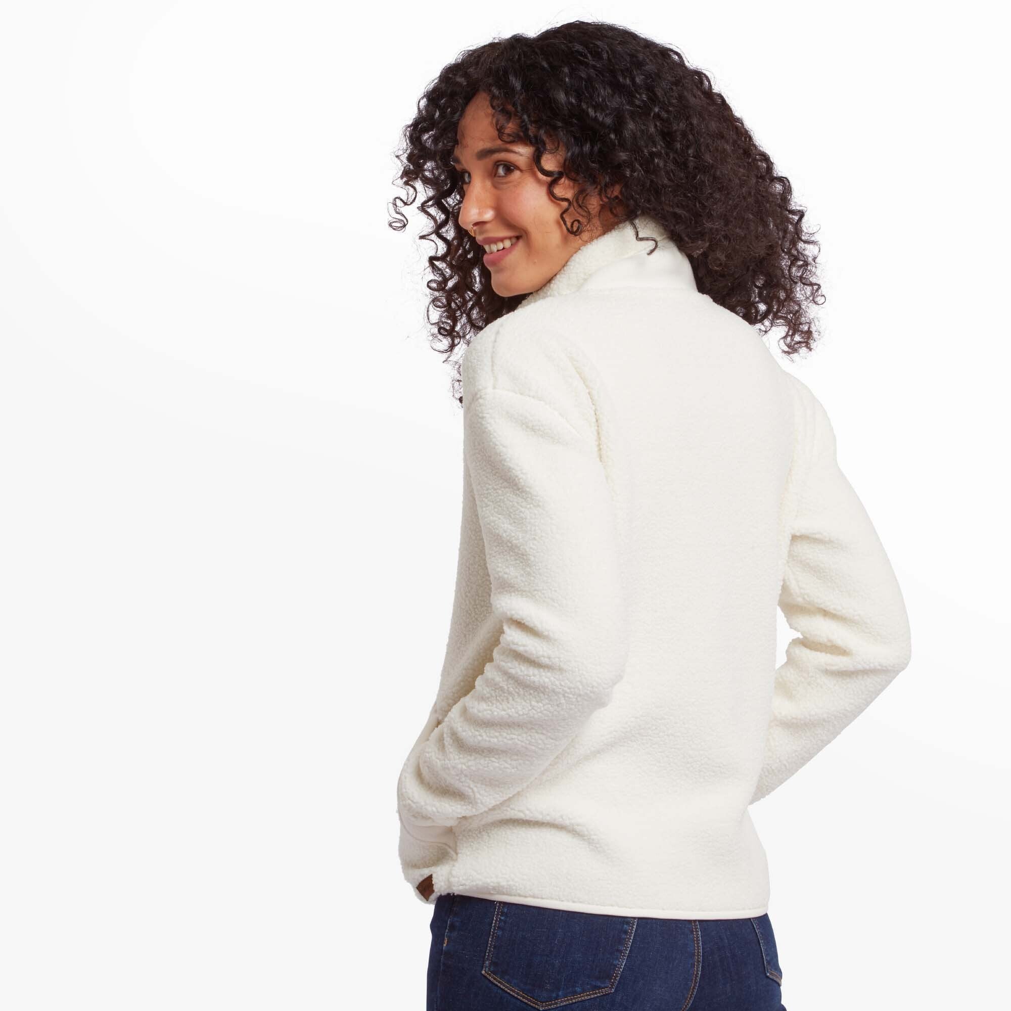 A back view of the Sherpa Adventure Gear Chamlang Full Zip Jacket in Cream. The high collar and soft texture of the fleece are highlighted. The woman has her hair styled in natural curls, and the jacket pairs seamlessly with her dark blue jeans.