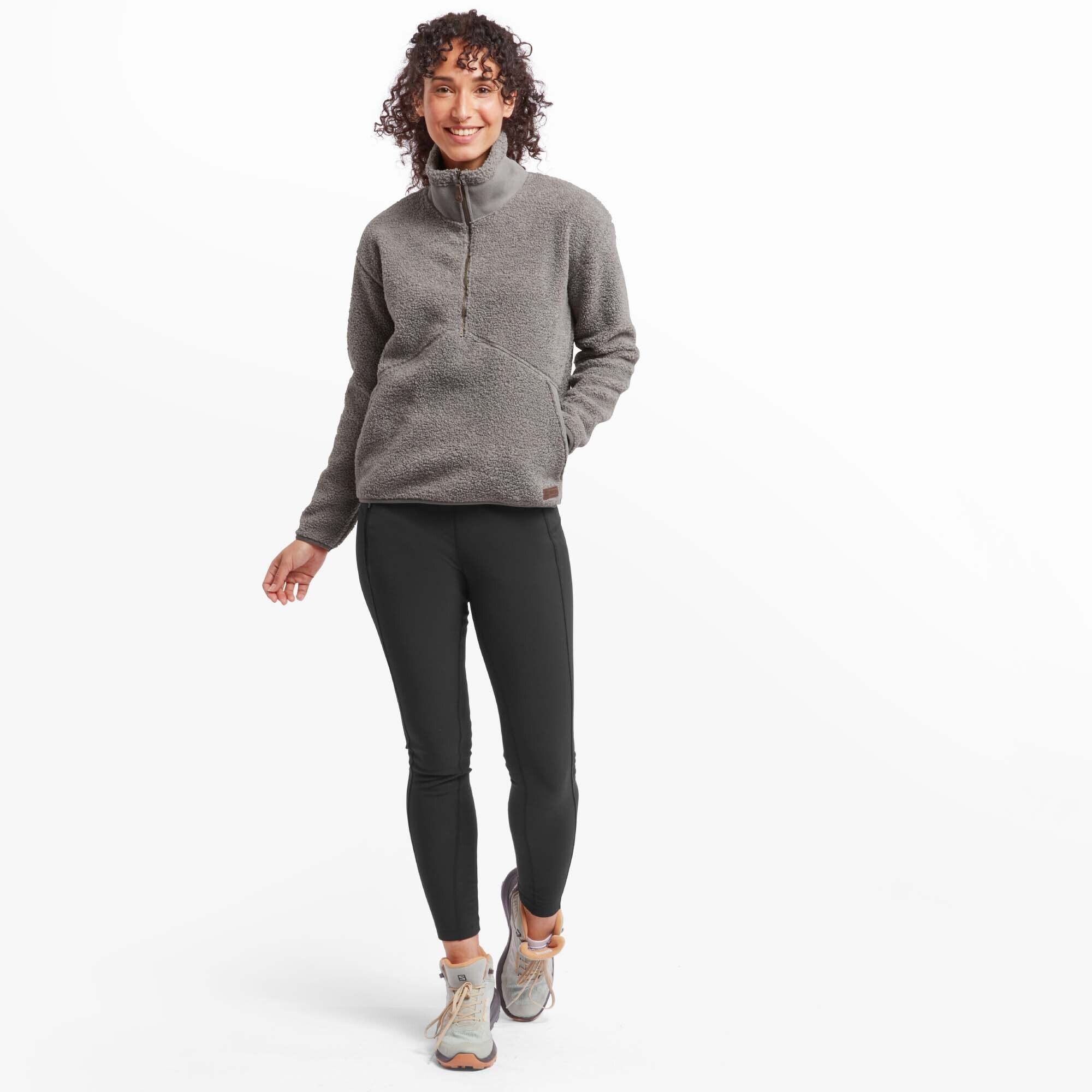 A full-body view of a woman wearing the Sherpa Adventure Gear Chamlang Half Zip Pullover in Grey with black leggings and grey hiking shoes. She is smiling warmly and standing casually, showcasing the pullover's relaxed fit and suitability for outdoor activities.