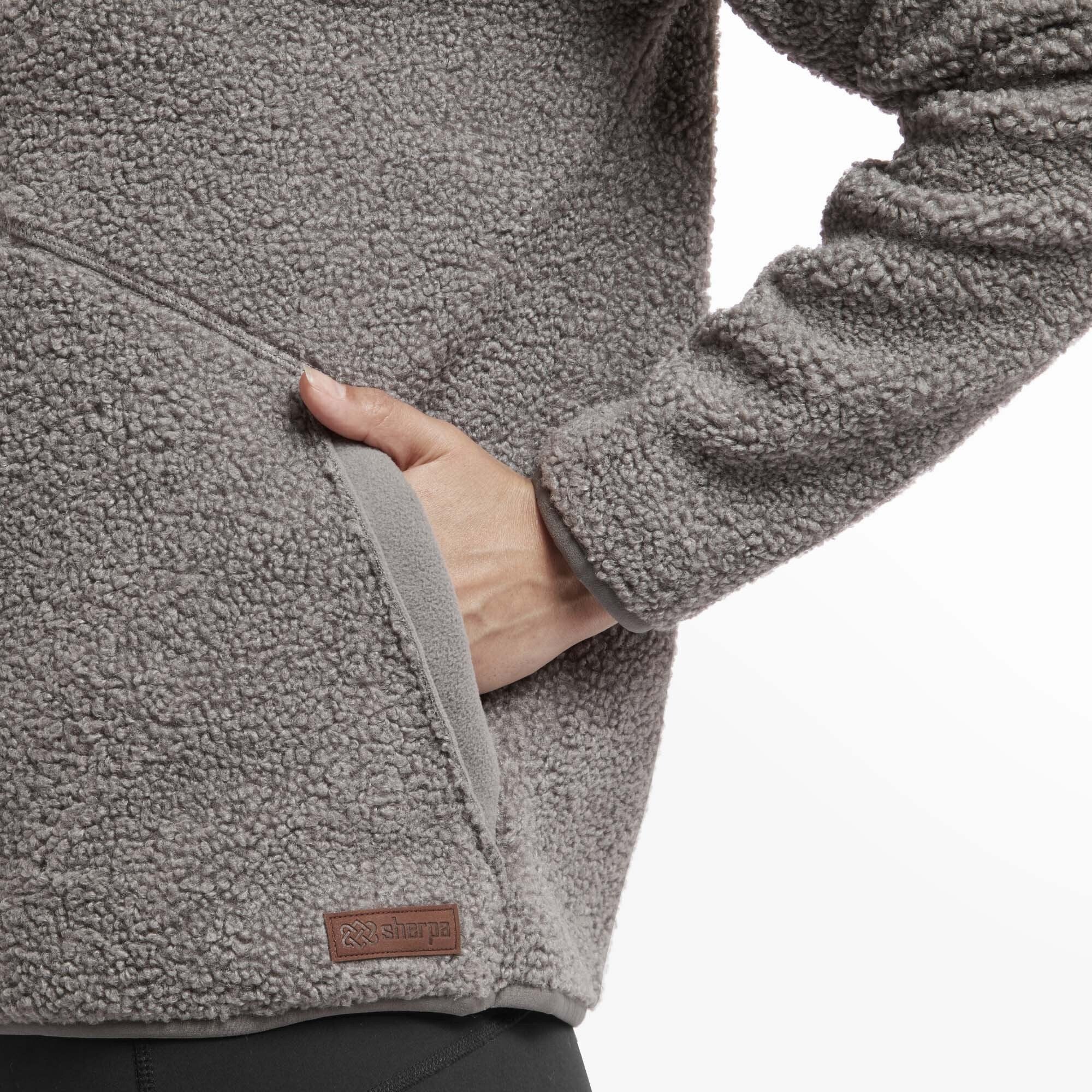 A close-up of the hand pocket on a Sherpa Adventure Gear Chamlang Half Zip Pullover in Grey. The texture of the thick, cozy fleece is highlighted, and a small brown Sherpa Adventure Gear logo tag is visible near the hem. The functional pocket design emphasises warmth and practicality.