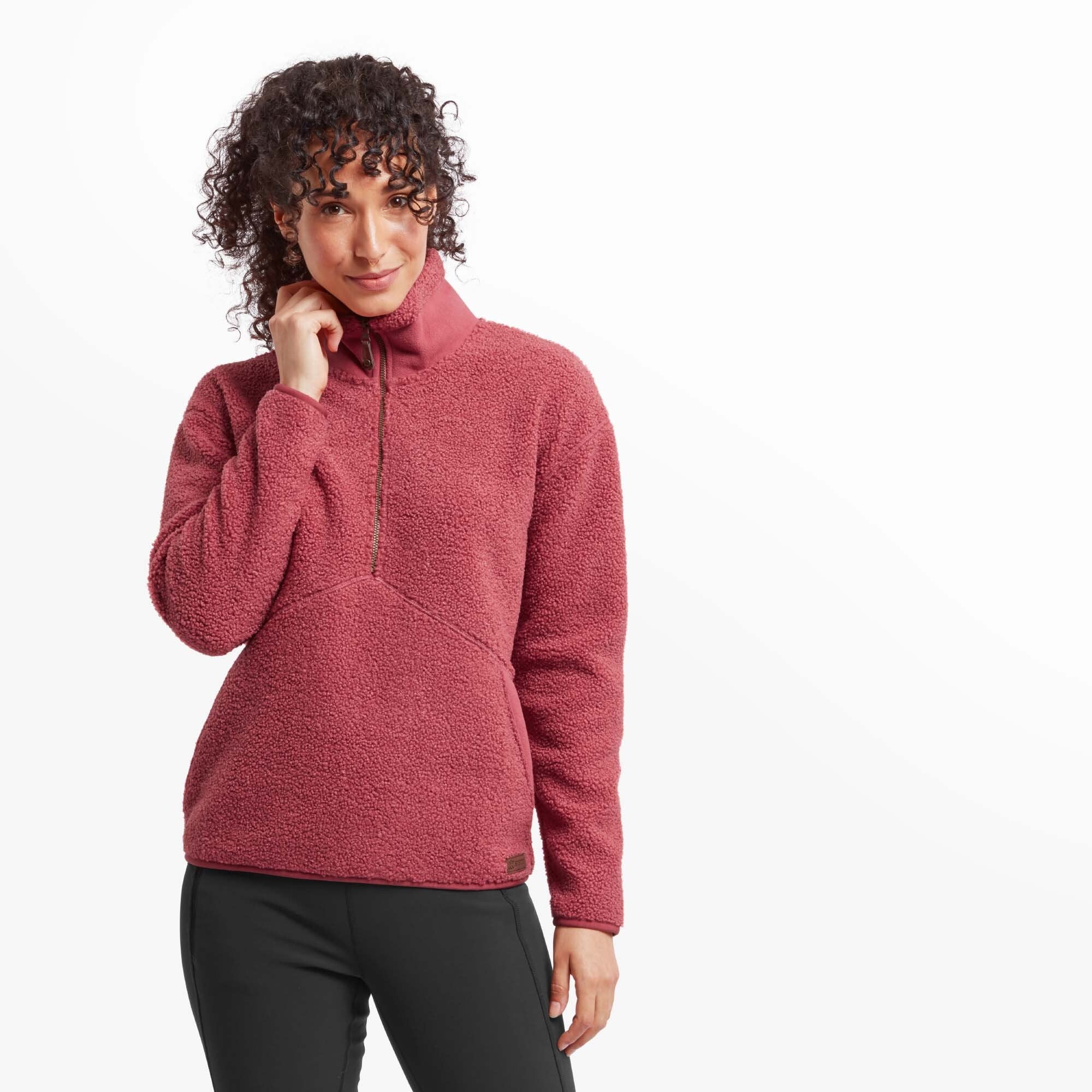 A front-facing view of a woman wearing a cozy Sherpa Adventure Gear Chamlang Half Zip Pullover in Red made of textured fleece. She is gently touching her chin, showcasing the high collar and the soft material of the pullover.