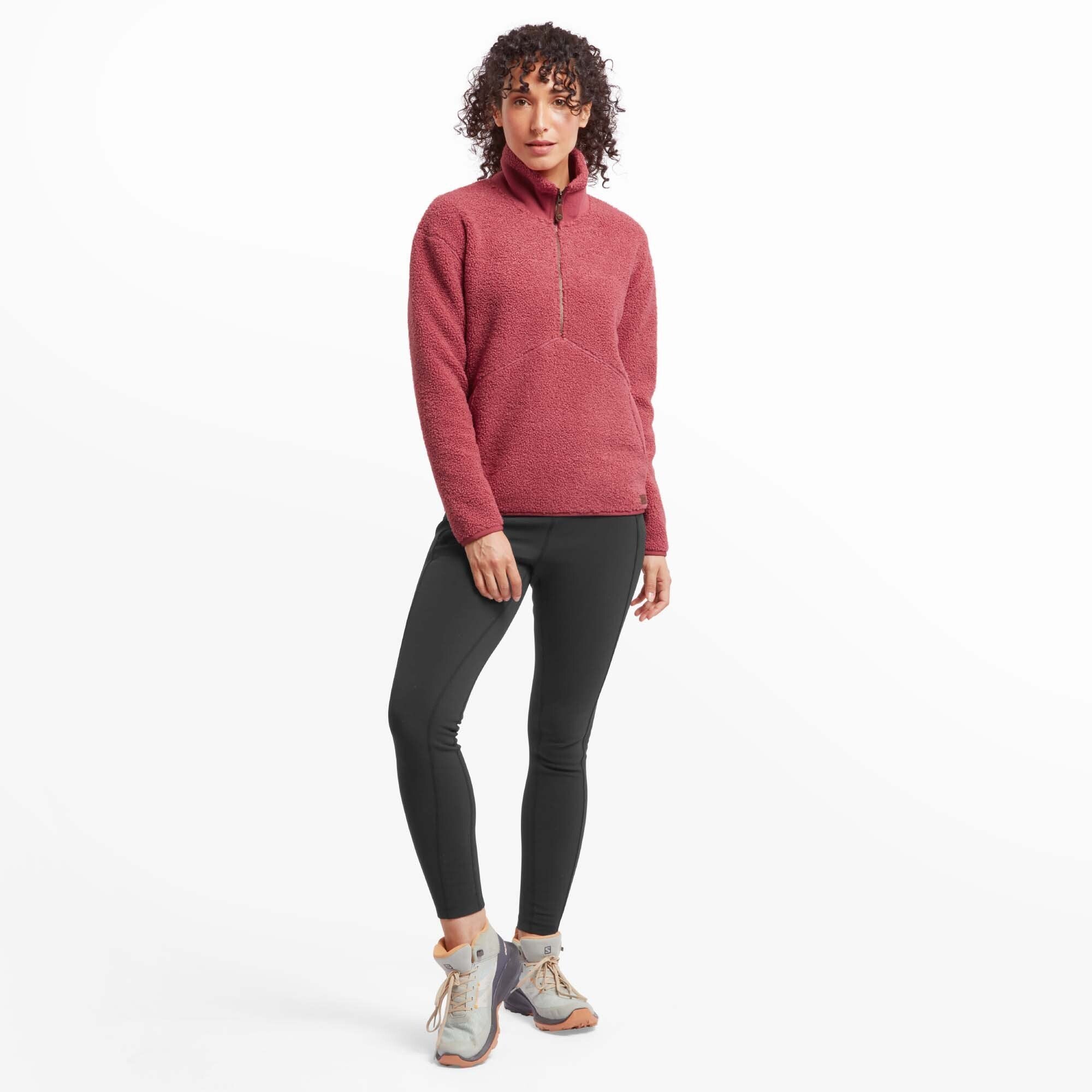 A full-body shot of the woman wearing the Sherpa Adventure Gear Chamlang Half Zip Pullover in Red with black leggings and light hiking shoes. She stands with her legs slightly apart, highlighting the casual yet outdoors-ready look of the outfit.