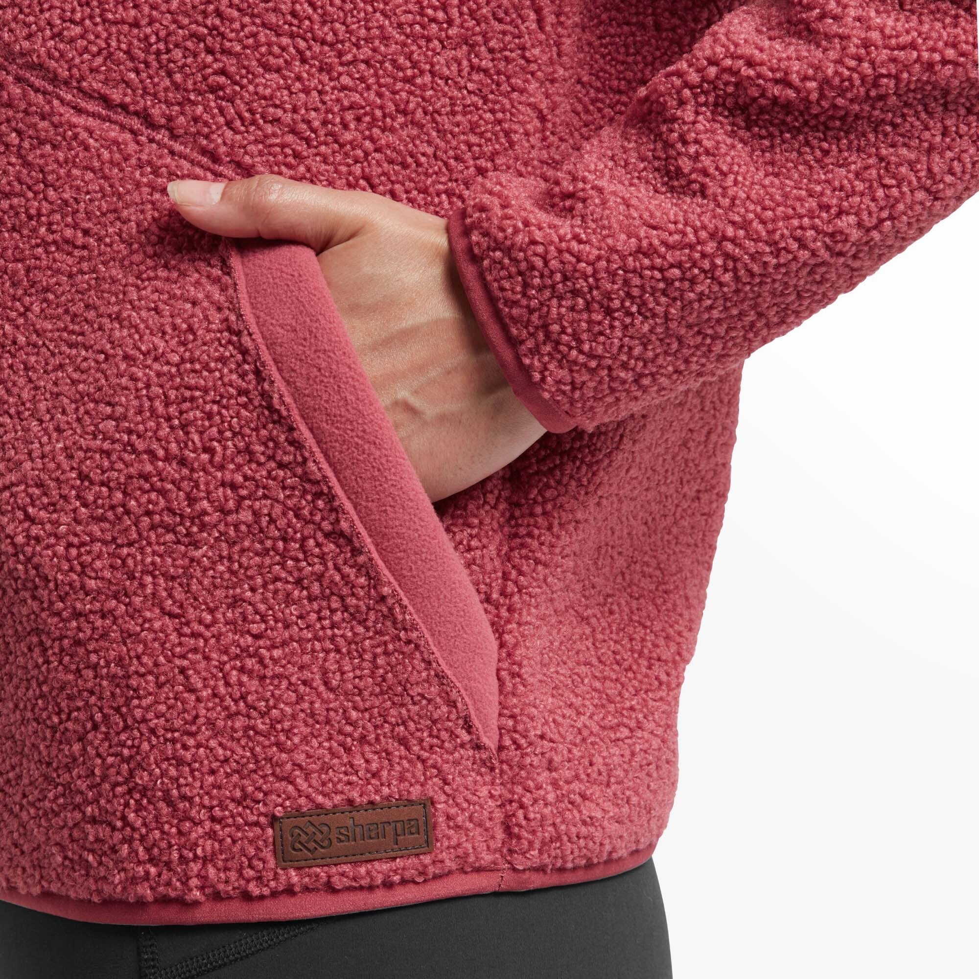 A detailed shot of the side pocket on the Sherpa Adventure Gear Chamlang Half Zip Pullover in Red. The pocket features a small branded label with the "Sherpa" logo and a fleece-lined interior for warmth and comfort.