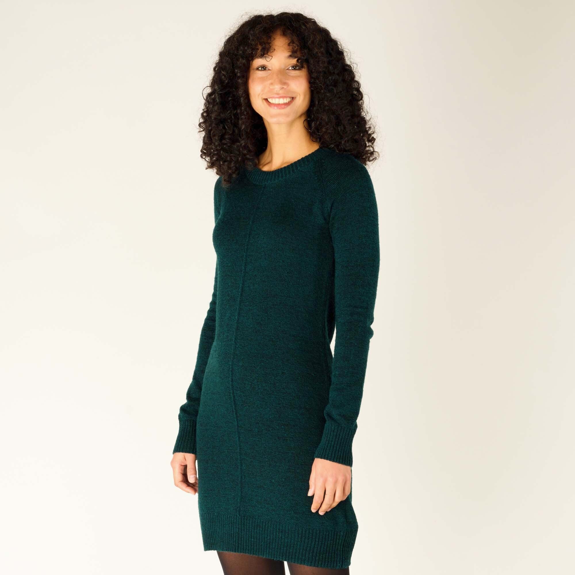 A female model with curly dark hair stands against a neutral background, smiling gently while wearing a Sherpa Adventure Gear Chimee Dress in Green. The dress is form-fitting with ribbed cuffs and hem, and a central seam running vertically down the front. The model's left arm hangs naturally at her side, while her right hand rests slightly in front of her. She pairs the dress with black tights.