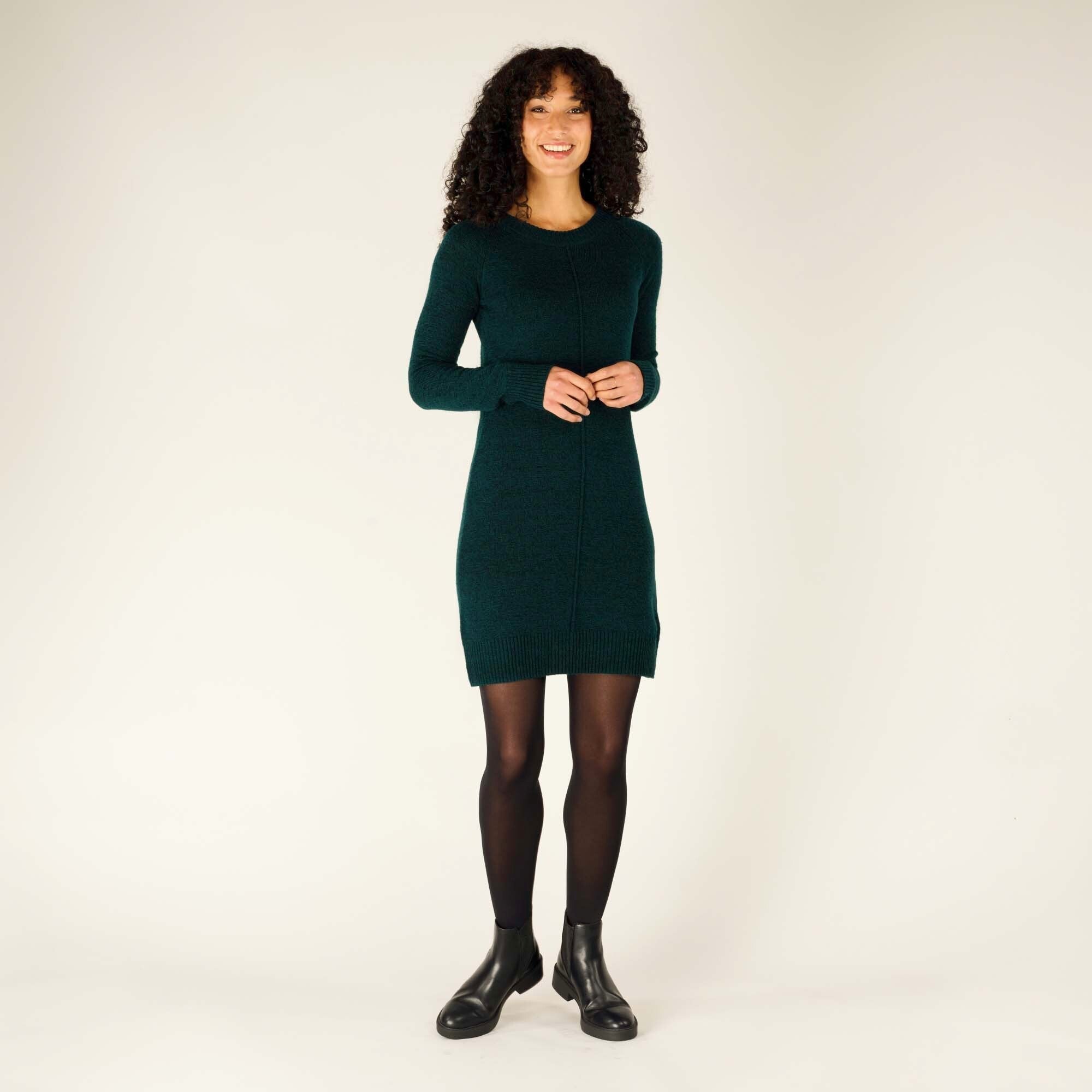 The same model poses with her feet slightly apart, hands clasped in front of her, showing the full view of the Sherpa Adventure Gear Chimee Dress in Green. The dress reaches just above the knees, and the fit highlights her natural shape. She completes the look with opaque black tights and black ankle boots with a modest heel.