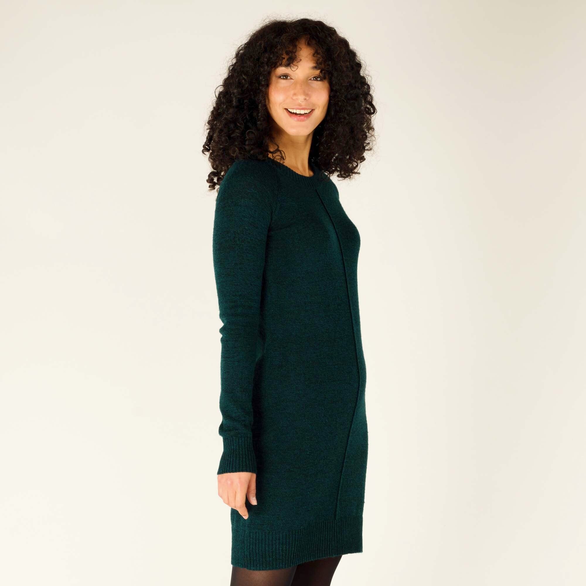 The model turns to her left, giving a profile view of the Sherpa Adventure Gear Chimee Dress in Green. The slim fit and soft knit fabric contour her silhouette. The ribbed detailing at the hem and cuffs is visible, as is the dress’s slight stretch. The model smiles warmly at the camera.