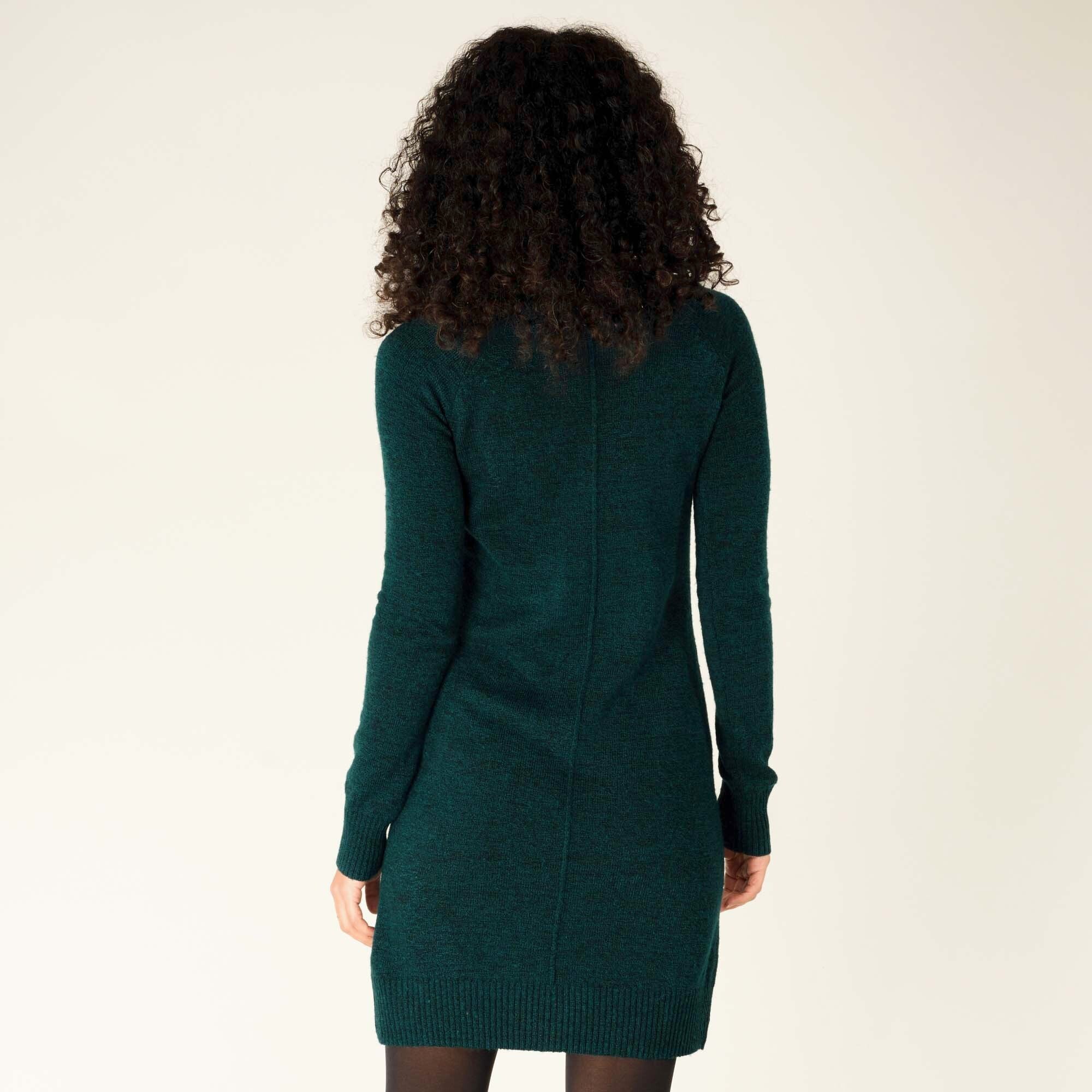 In this shot, the model stands with her back to the camera, showing the rear design of the Sherpa Adventure Gear Chimee Dress in Green. A central seam runs vertically down the back, and the ribbed hem hugs her legs slightly above the knee. Her curly hair frames her upper back.