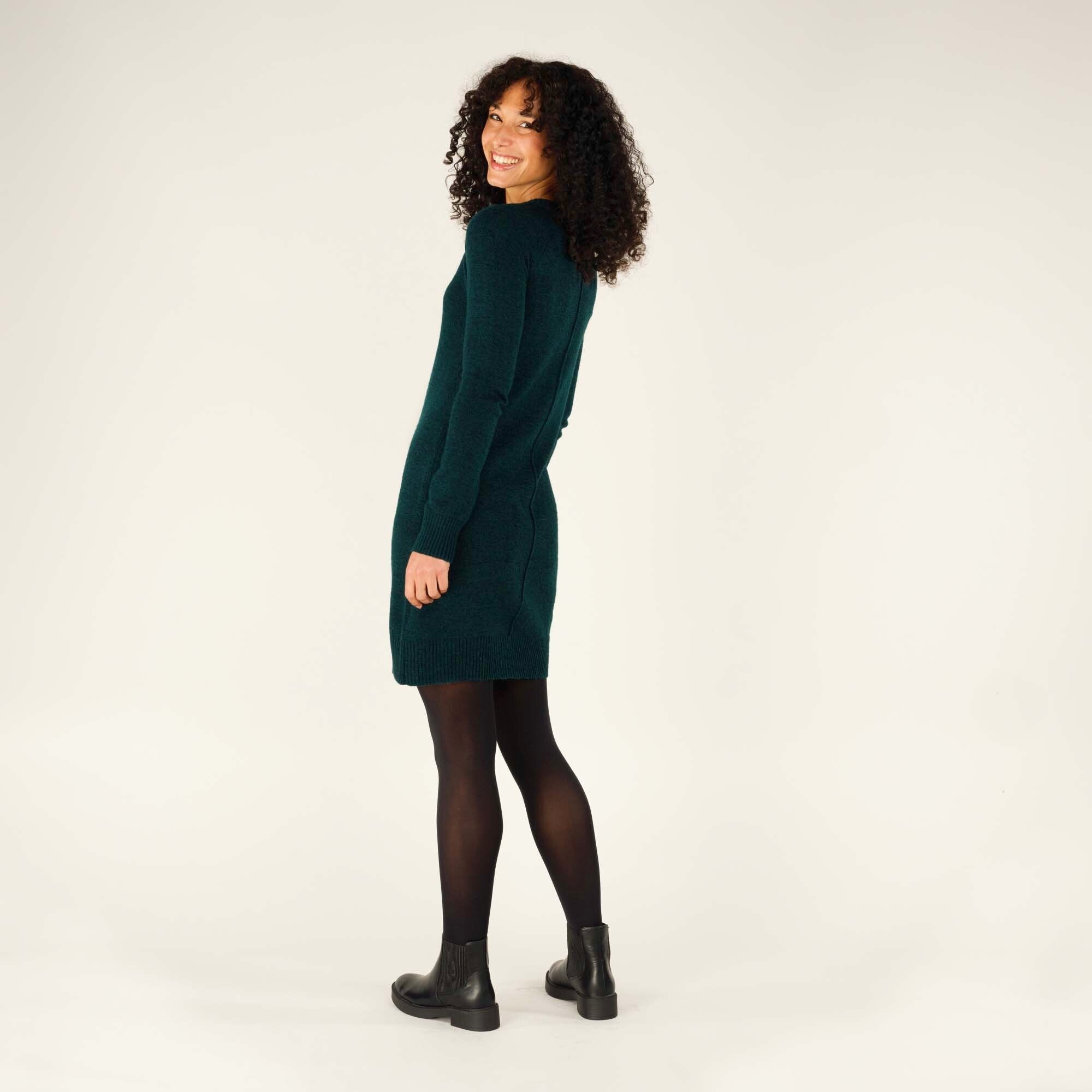 The model turns slightly to her right, glancing back over her shoulder with a smile. This angle showcases both the back and side of the Sherpa Adventure Gear Chimee Dress in Green, highlighting the soft drape of the knit fabric and the flattering fit. Her black tights and boots complete the outfit.