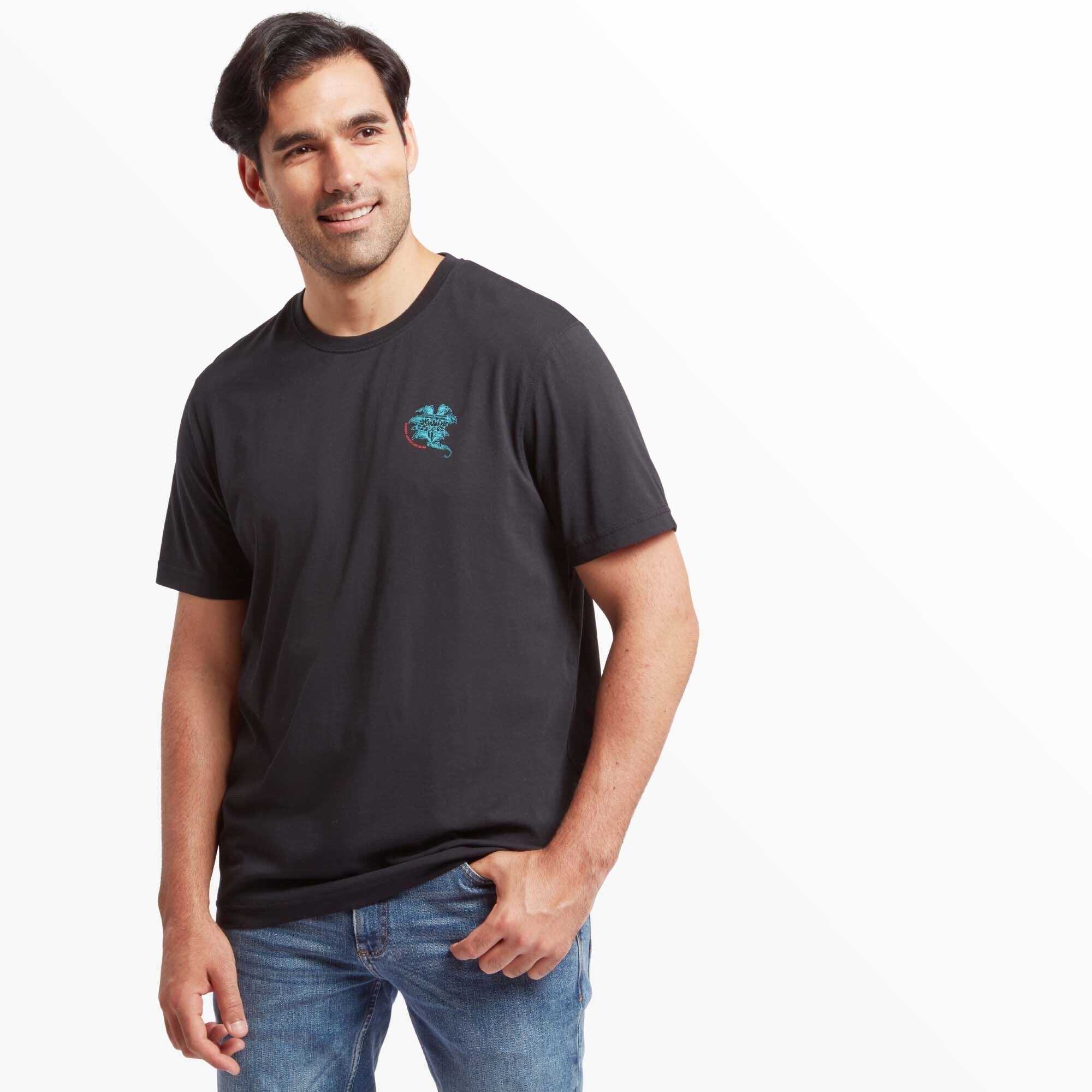 A man modeling the Sherpa Adventure Gear Deity T-Shirt in Black, standing casually with one hand in his pocket. The T-shirt features the same lotus flower design in turquoise and red on the chest. He pairs the shirt with light blue jeans and brown boots.