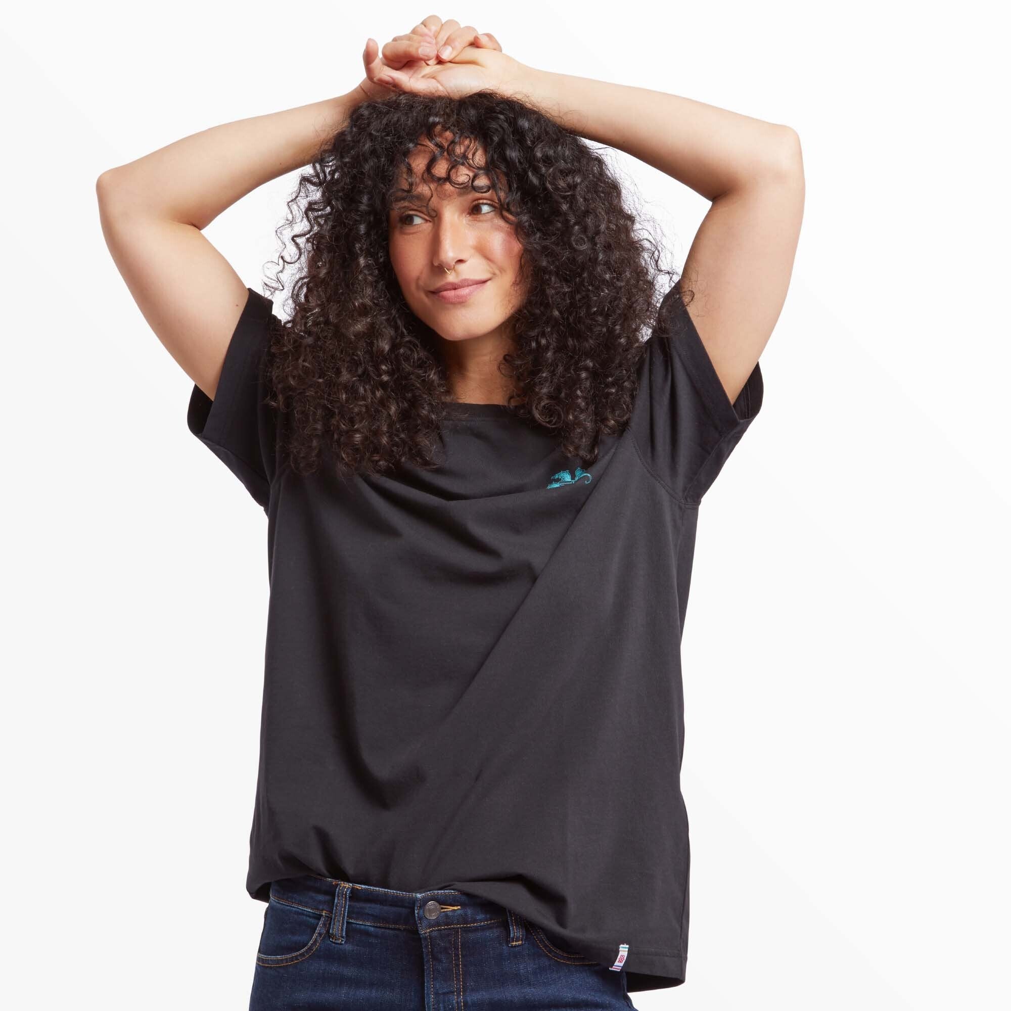 A woman modeling the Sherpa Adventure Gear Deity T-Shirt in Black, styled as an oversized fit with rolled-up short sleeves. She poses with her arms raised above her head, showcasing the relaxed and casual design. The lotus flower design is visible on the chest, paired with dark denim jeans.