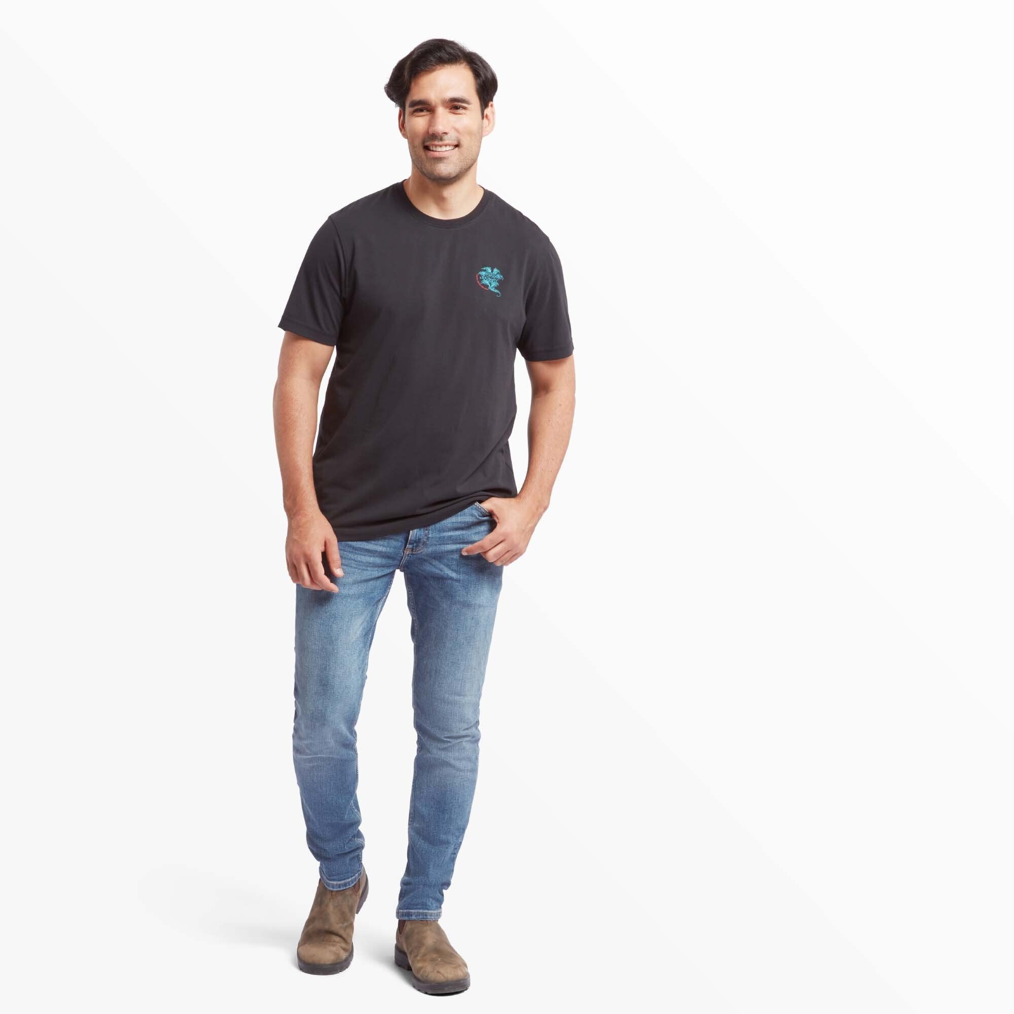 A full-length view of a man in the Sherpa Adventure Gear Deity T-Shirt in Black, styled with light blue jeans and brown boots. The man stands confidently with a smile, highlighting the shirt’s comfortable fit and versatile style.