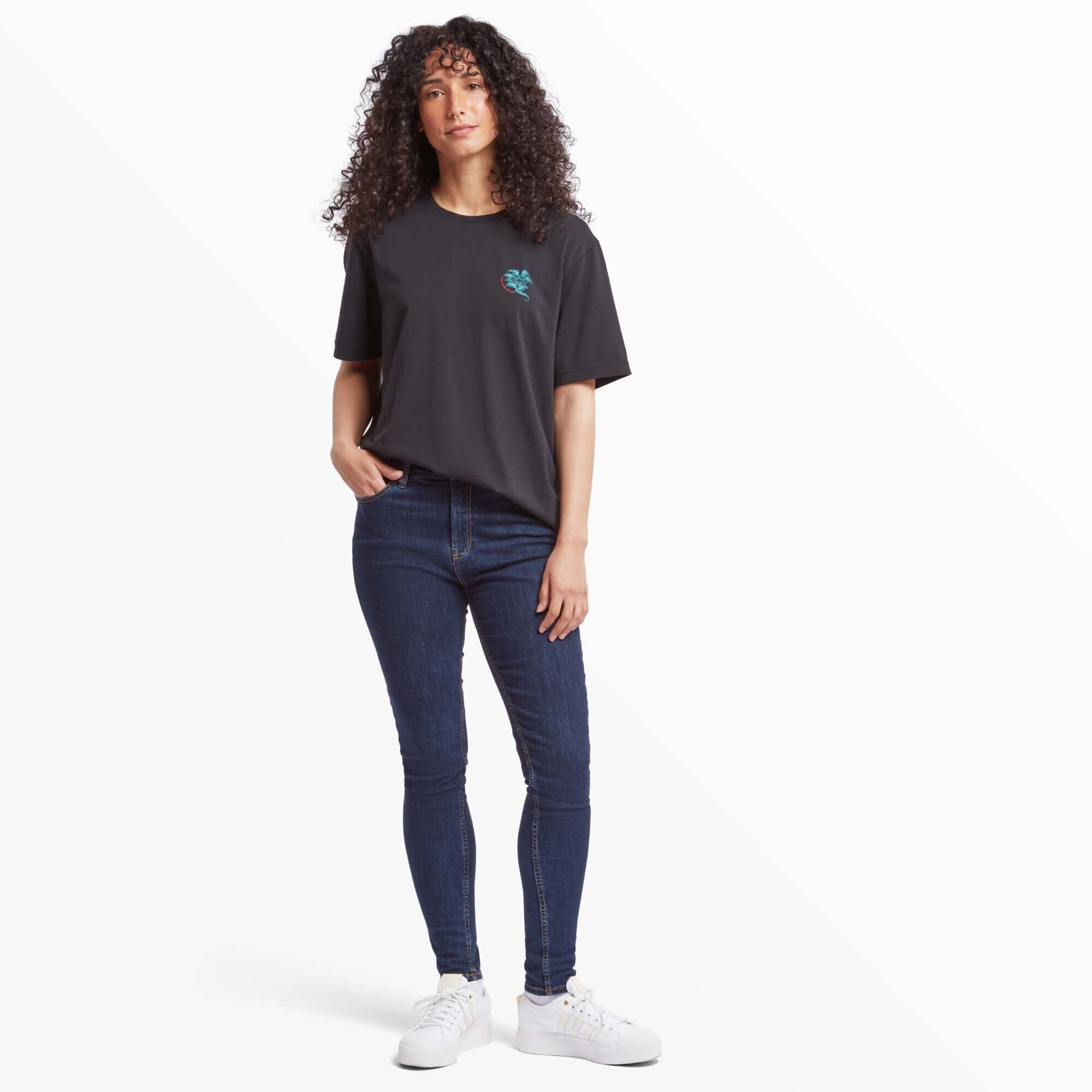 A full-length view of a woman wearing the Sherpa Adventure Gear Deity T-Shirt in Black paired with skinny dark denim jeans and white trainers. She stands with one hand in her pocket, showcasing a relaxed yet polished look.