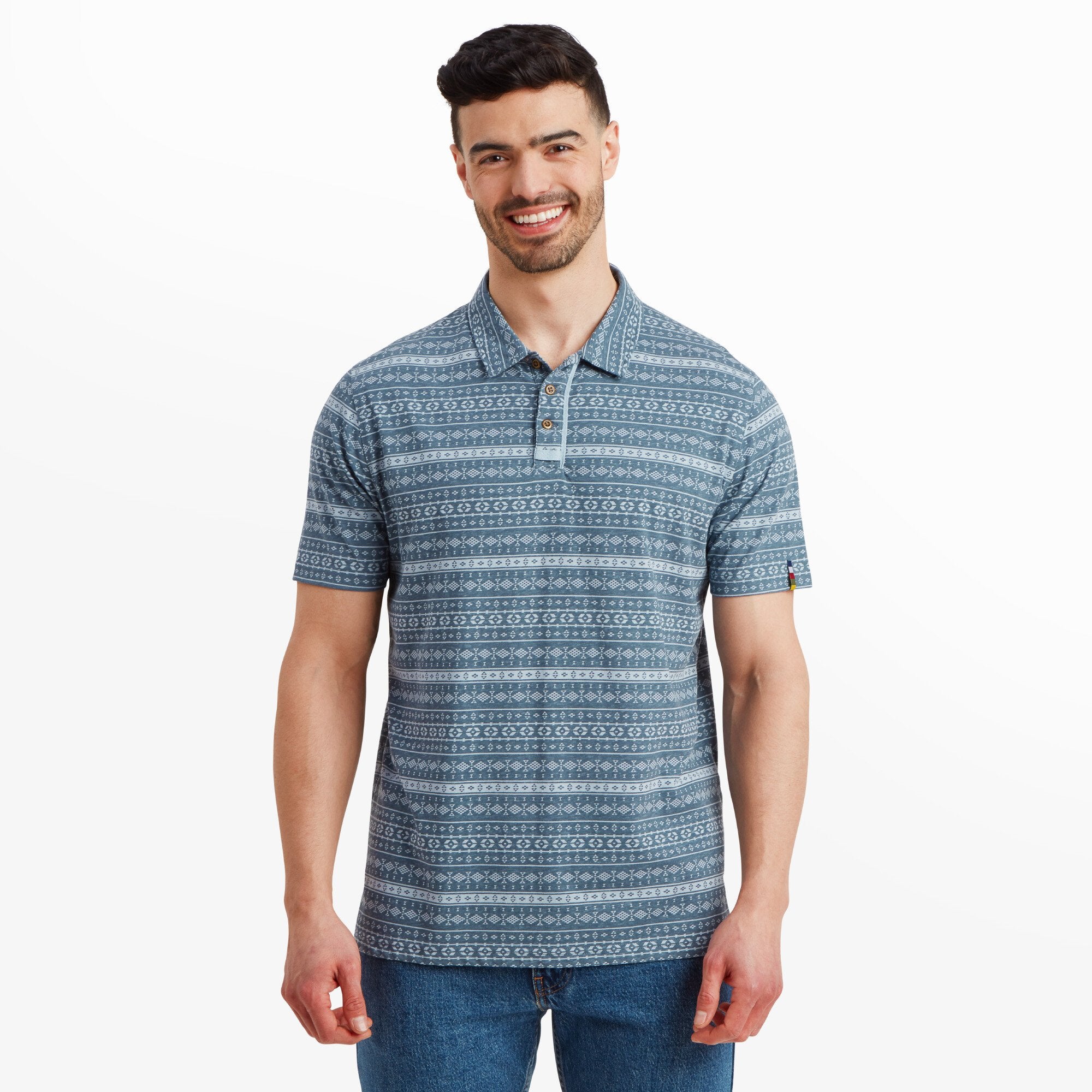 A man is wearing a Sherpa Adventure Gear Dhani Polo Shirt in Blue with a subtle geometric pattern featuring stripes and small diamond shapes. The shirt has a classic collar with a three-button placket. He is smiling and standing casually, paired with blue jeans.