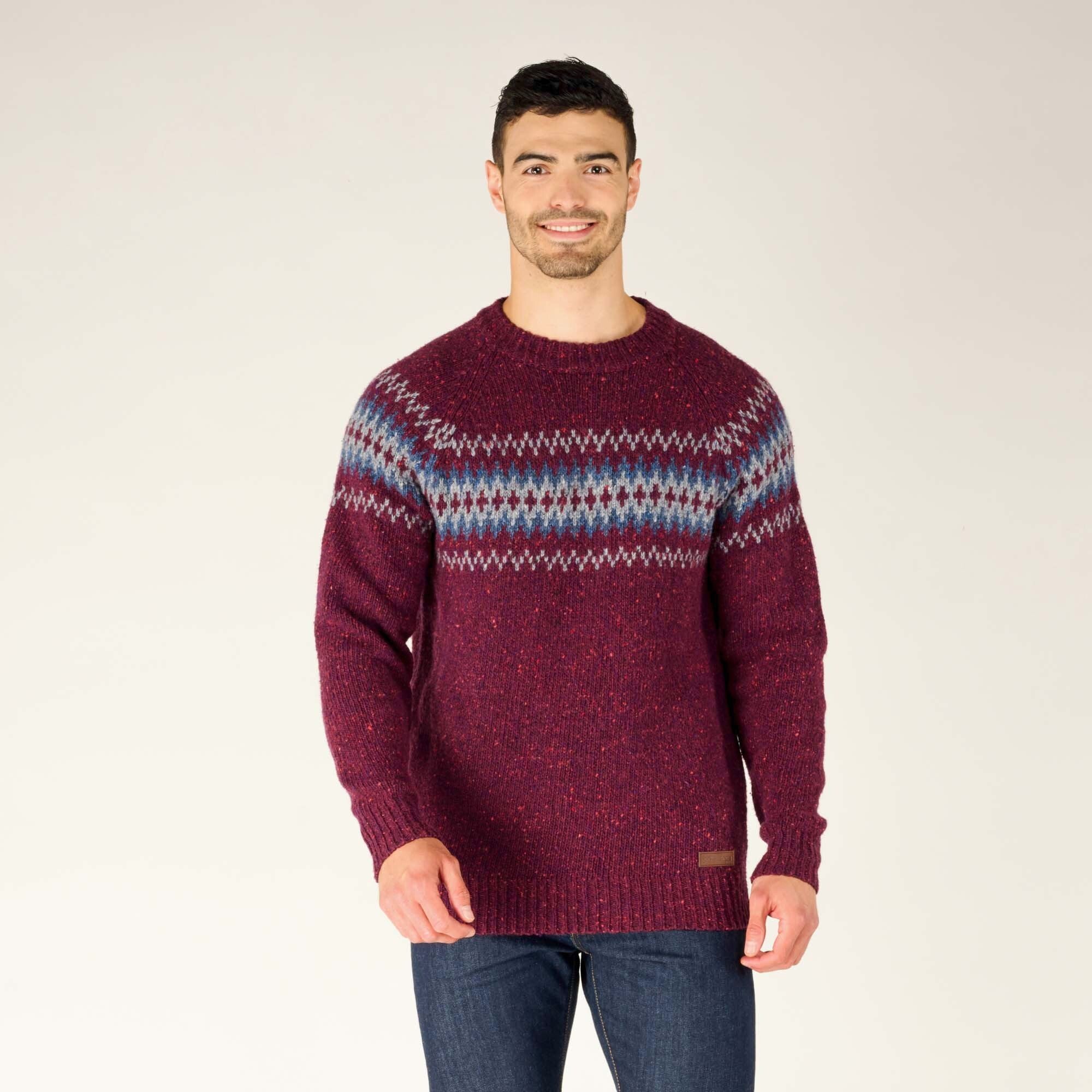 The man is smiling directly at the camera while wearing the Sherpa Adventure Gear Dumji Sweater in Red. This view clearly displays the vibrant fair-isle pattern and the rich speckled texture of the knit material. The sweater has a ribbed hem and cuffs, and he keeps his hands relaxed at his sides.