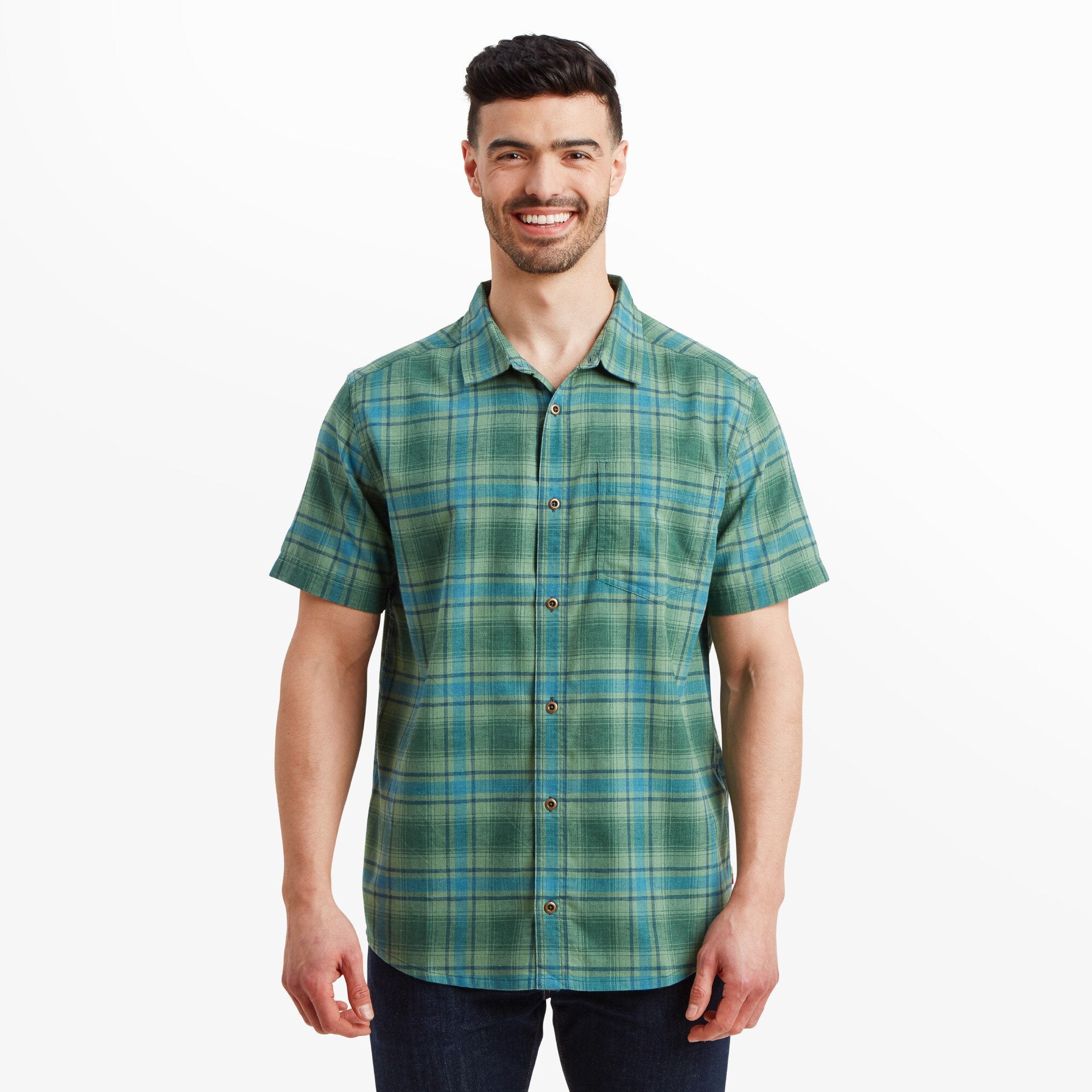 A man wearing a Sherpa Adventure Gear Faneel Plaid Short Sleeve Shirt in Green, standing and smiling confidently. The shirt features a classic button-down style with a left chest pocket, paired with dark blue jeans. The fabric showcases a vibrant green base with blue plaid patterns.