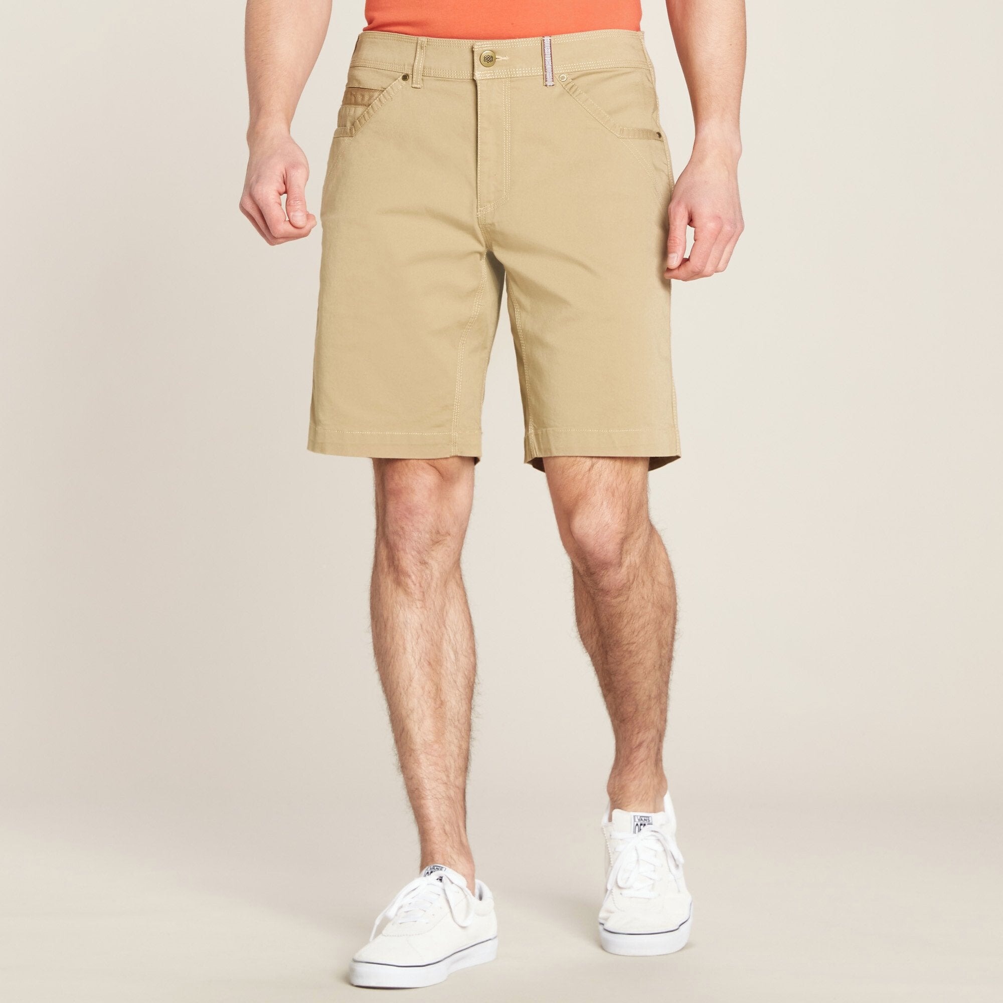 A male model wearing Sherpa Adventure Gear Guide Shorts in Beige from the back stands facing the camera. The shorts have a classic fit, falling just above the knee, with front pockets on each side. The waistband features a button closure and a zipper fly, along with a decorative woven belt loop detail. The model pairs the shorts with an orange shirt tucked in and white casual sneakers. The background is a neutral beige tone.