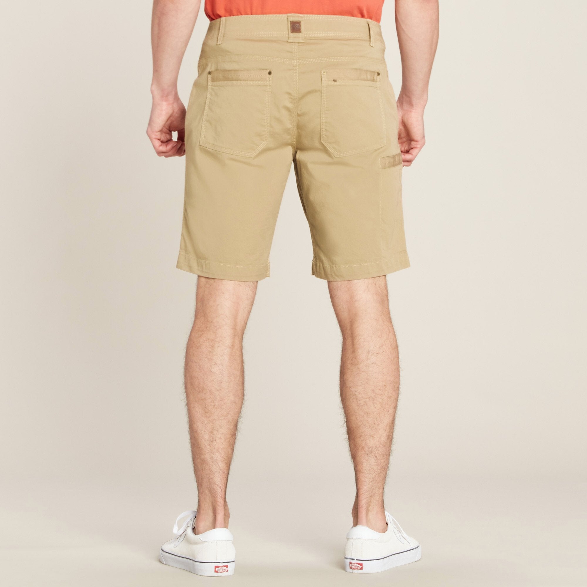 The same male model stands with his back facing the camera, wearing the Sherpa Adventure Gear Guide Shorts in Beige from the back. The rear view showcases two back pockets with button closures and reinforced stitching. There is also a small tool pocket on the right side. A small Sherpa Adventure Gear logo patch is visible on the back waistband. The shorts have a relaxed fit and durable fabric. The model’s legs are slightly apart, showing the shorts' movement and flexibility.