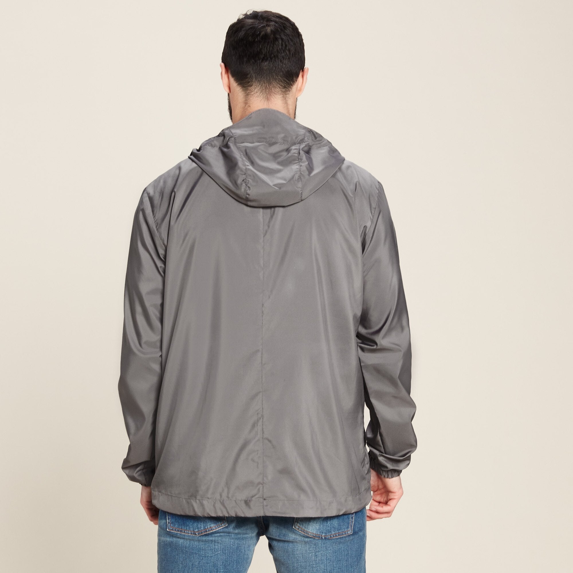 A rear view of the man wearing the Sherpa Adventure Gear Halka Anorak Jacket in Grey. The hood is visible in its relaxed state, and the back hem of the jacket extends slightly below the waist. The jacket's lightweight material and loose fit are emphasised. The man pairs the jacket with blue jeans, standing in a casual pose.