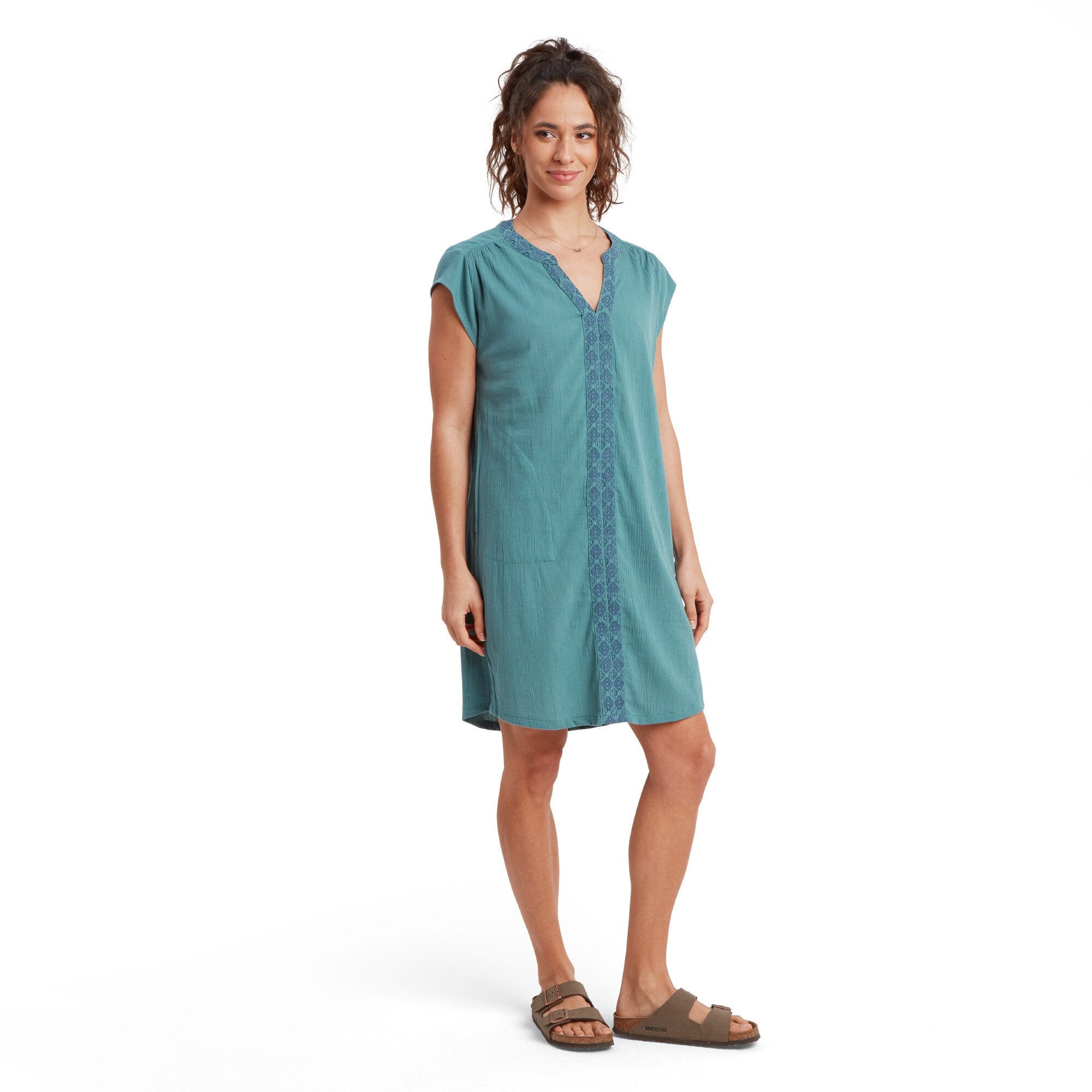 The image features a woman modeling a Sherpa Adventure Gear Hara Cap Sleeve Dress in Blue. The dress has a relaxed fit, falling just above the knees. A vertical embroidered pattern runs along the front of the dress, adding a touch of texture and detail. The neckline is a soft V-shape with intricate stitching along the edges. She pairs the dress with brown sandals.