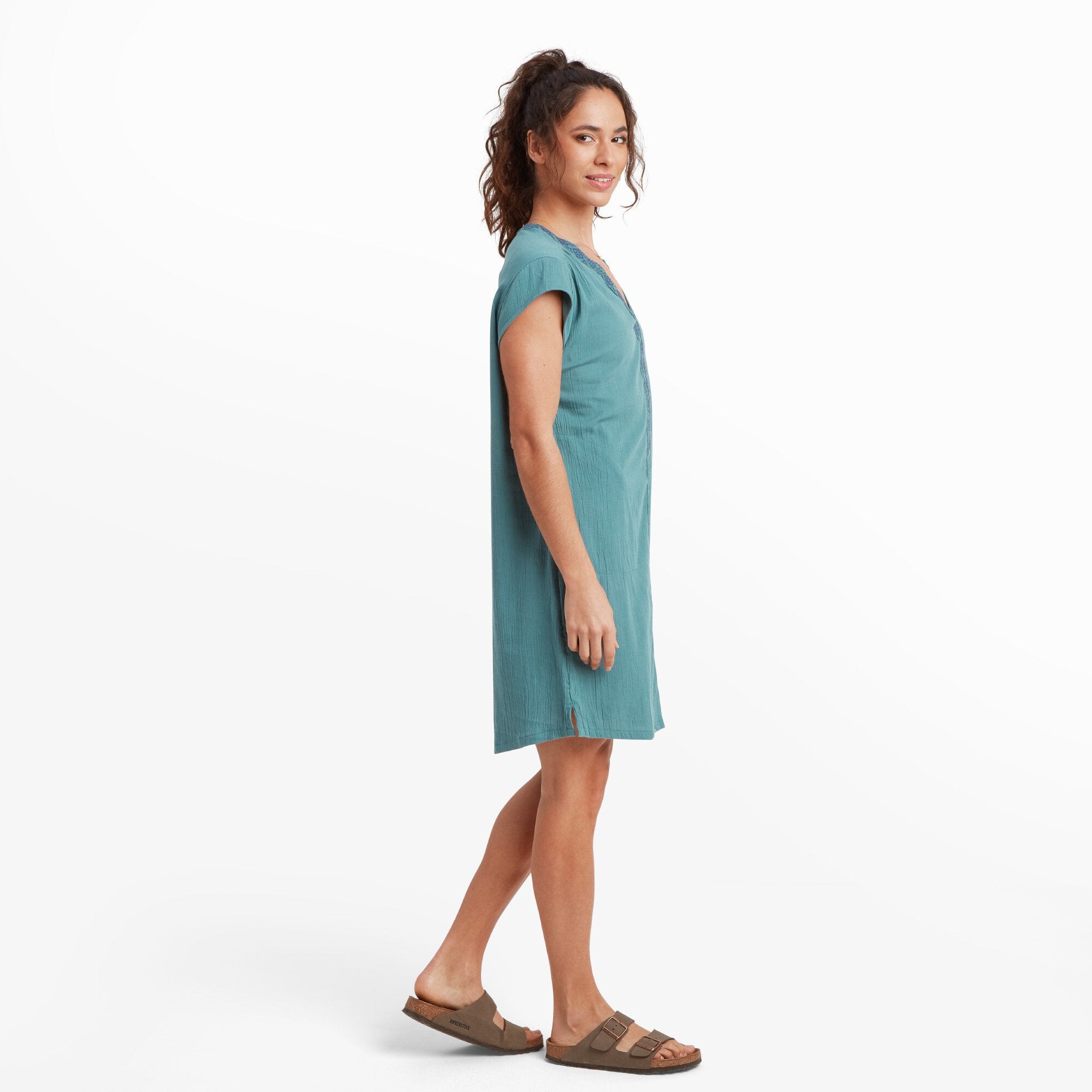 This image shows the woman from a side profile, emphasising the Sherpa Adventure Gear Hara Cap Sleeve Dress in Blue's loose and comfortable silhouette. The slight slit at the side hem is visible, providing ease of movement. The dress maintains its textured fabric and embroidered detailing along the front.