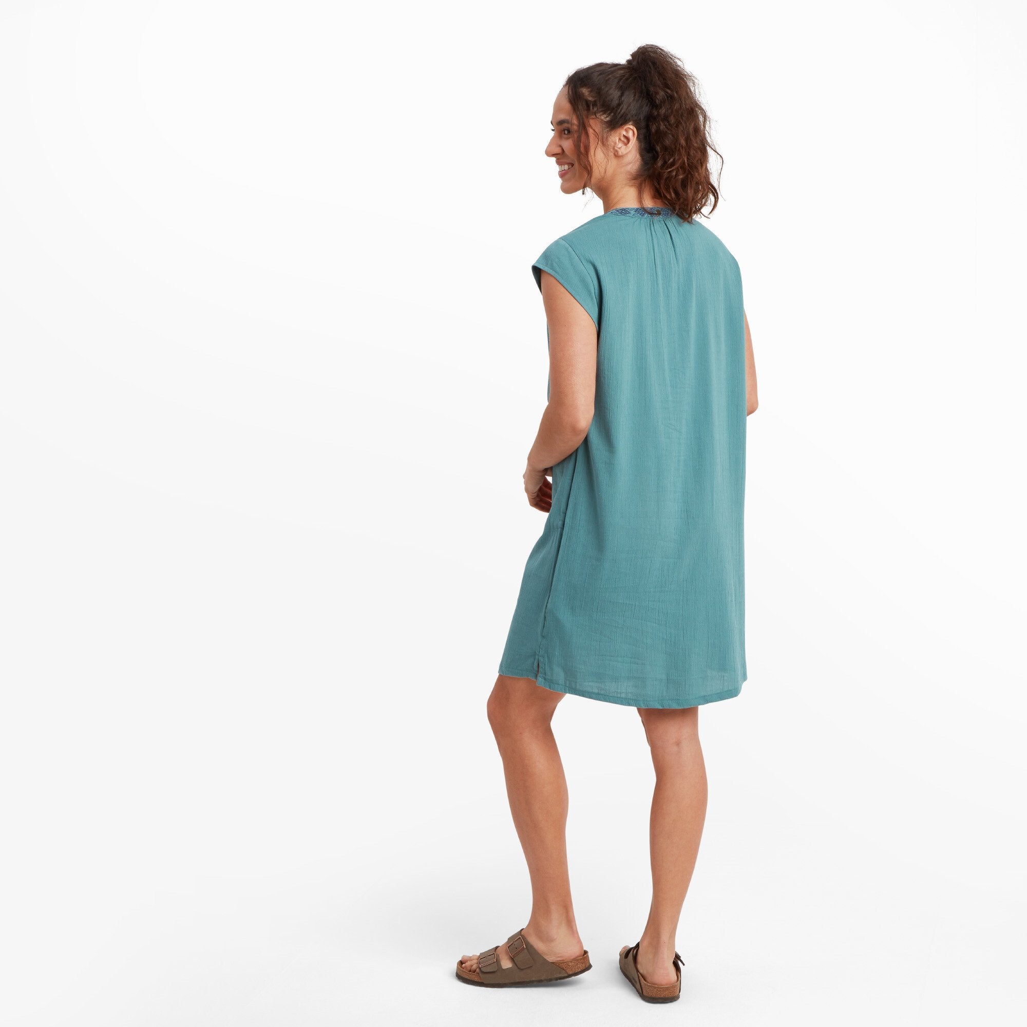 The model is shown from the back of the Sherpa Adventure Gear Hara Cap Sleeve Dress in Blue, showcasing the gathered detail at the top of the back neckline. A small logo embellishment is centered just below the neckline. The dress maintains its soft drape and flowy style.