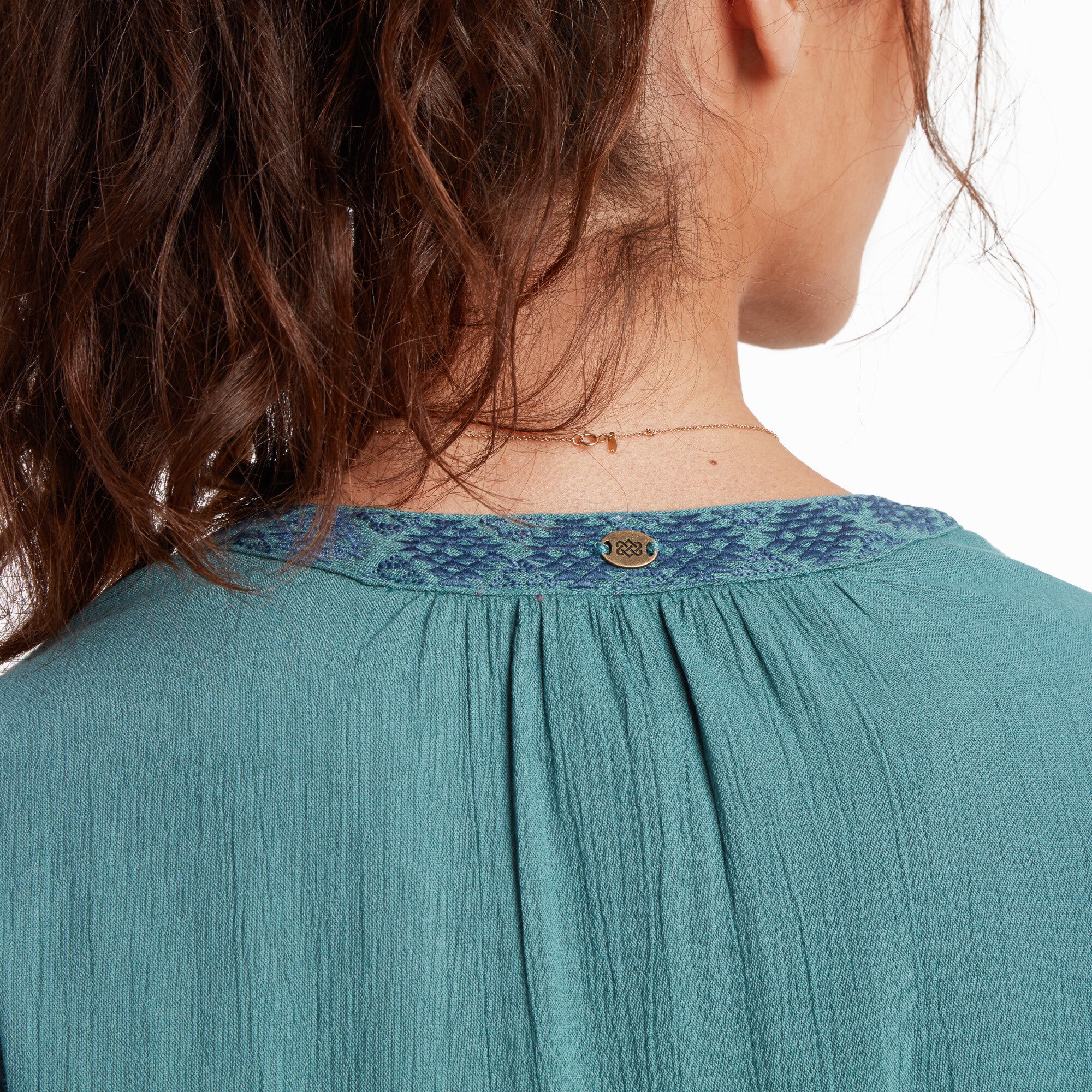 A close-up of the back neckline reveals the textured gathering at the top of the Sherpa Adventure Gear Hara Cap Sleeve Dress in Blue, accompanied by a small metallic logo embellishment that adds a subtle decorative touch. The stitching around the neckline is clearly visible, showcasing the craftsmanship.