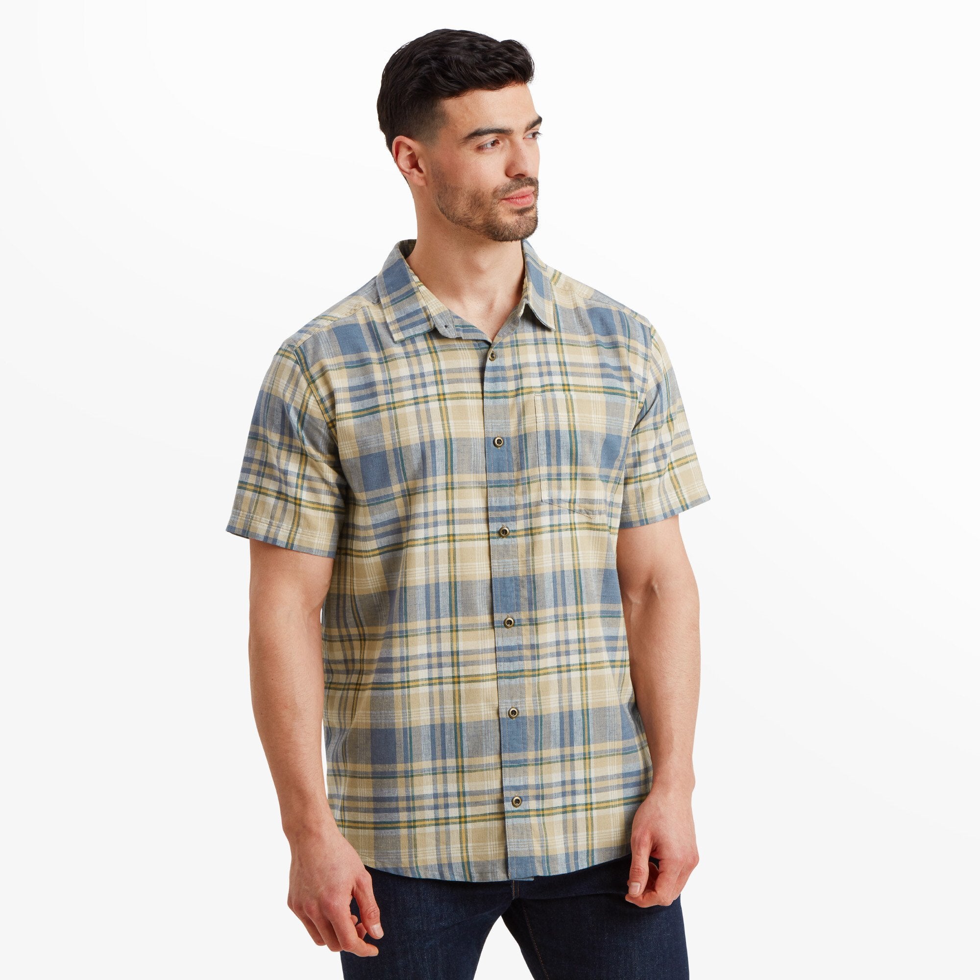 A man wearing a Sherpa Adventure Gear Hosh Plaid Short Sleeve Shirt in Beige, standing upright and facing slightly to his left. The shirt features a button-up front and a chest pocket. The model pairs the shirt with dark jeans, presenting a casual yet polished look.