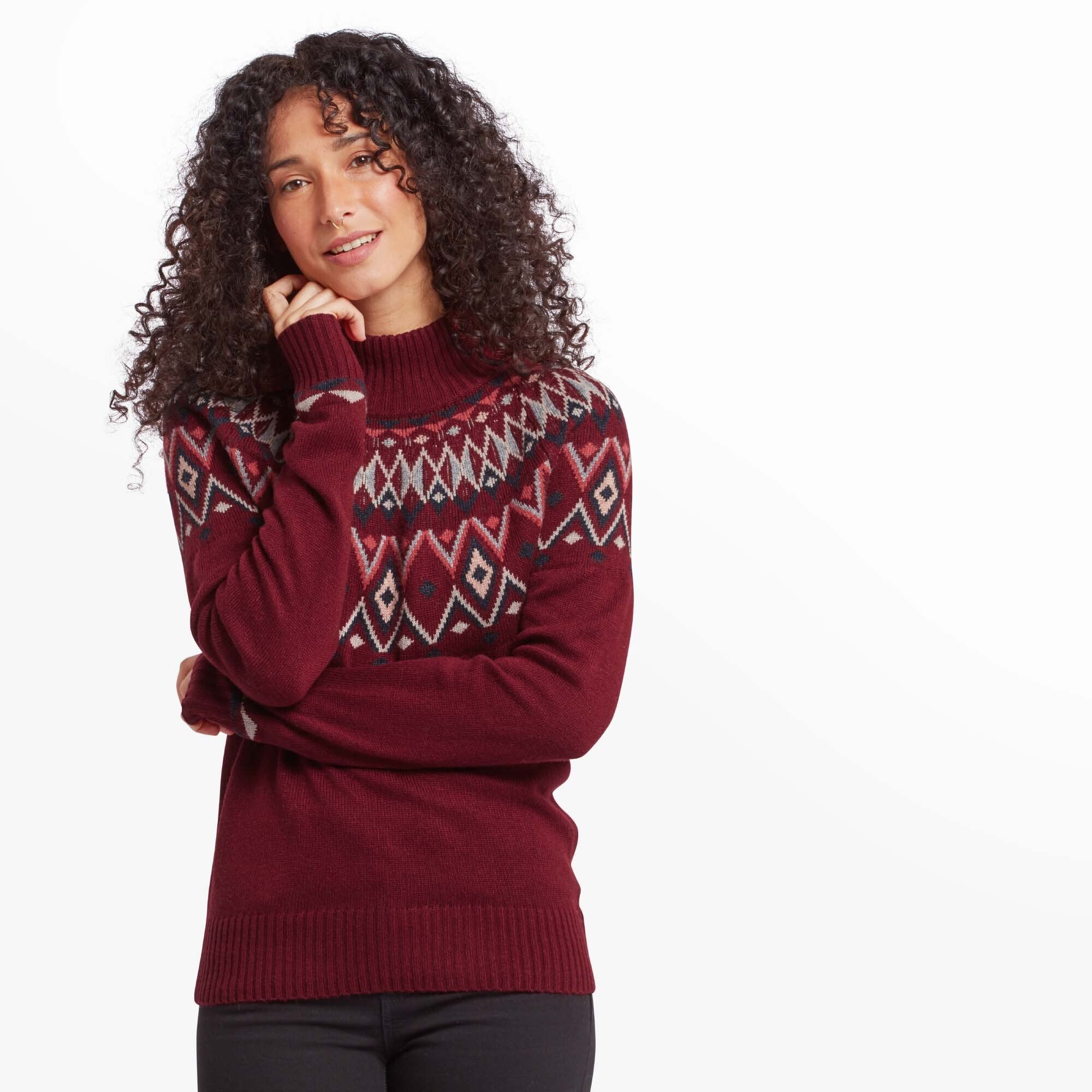 A woman with curly hair wearing a Sherpa Adventure Gear Indu Eco Mock Neck Jumper in Red featuring a geometric Fair Isle pattern in shades of cream, navy, and red. She is smiling gently, resting her hand on her cheek, with a clean white background emphasising the vibrant sweater.