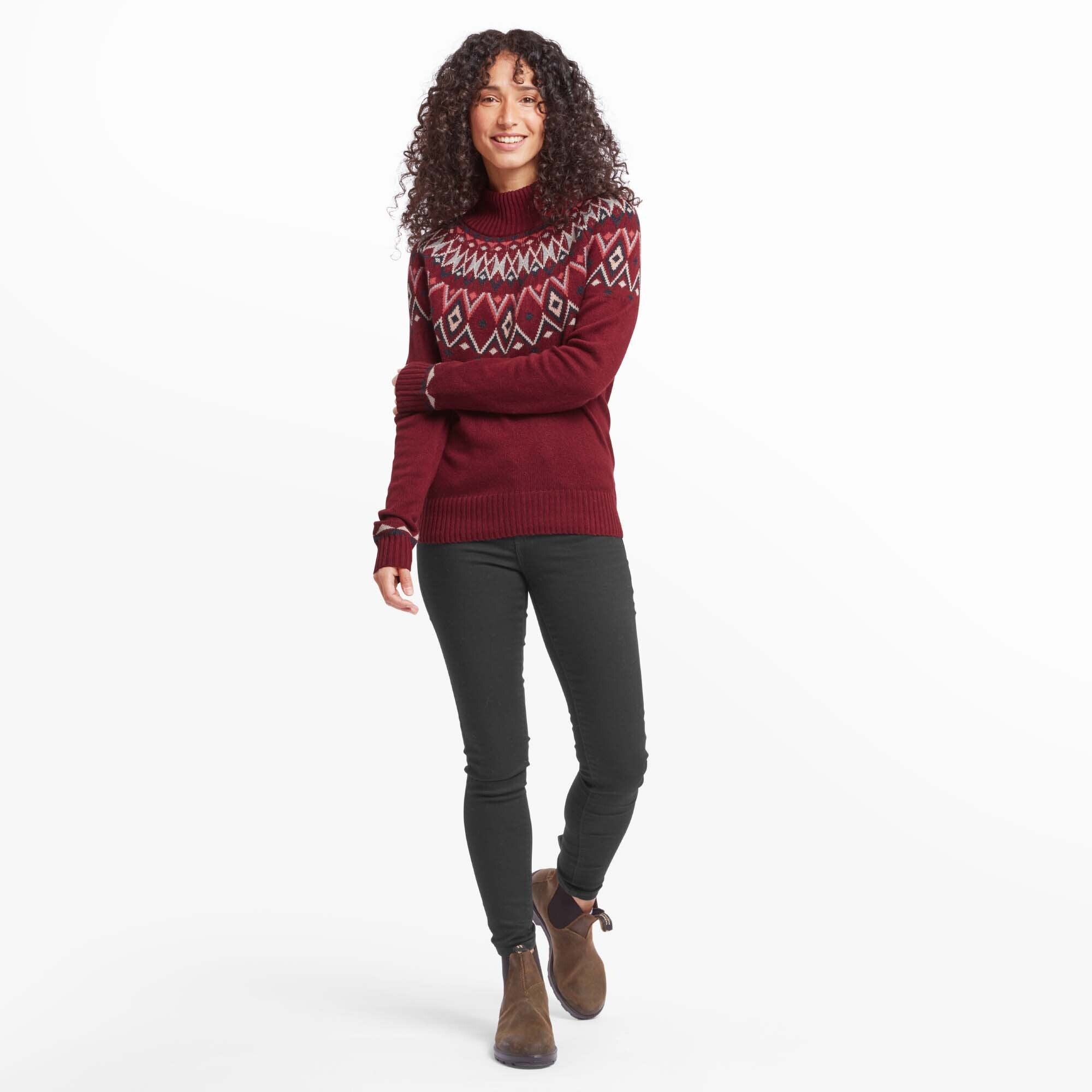 A full-body view of the woman wearing the Sherpa Adventure Gear Indu Eco Mock Neck Jumper in Red paired with black skinny jeans and brown ankle boots. She stands with a relaxed pose, slightly turned, while smiling warmly. The geometric pattern on the jumper adds a touch of style to her casual look.