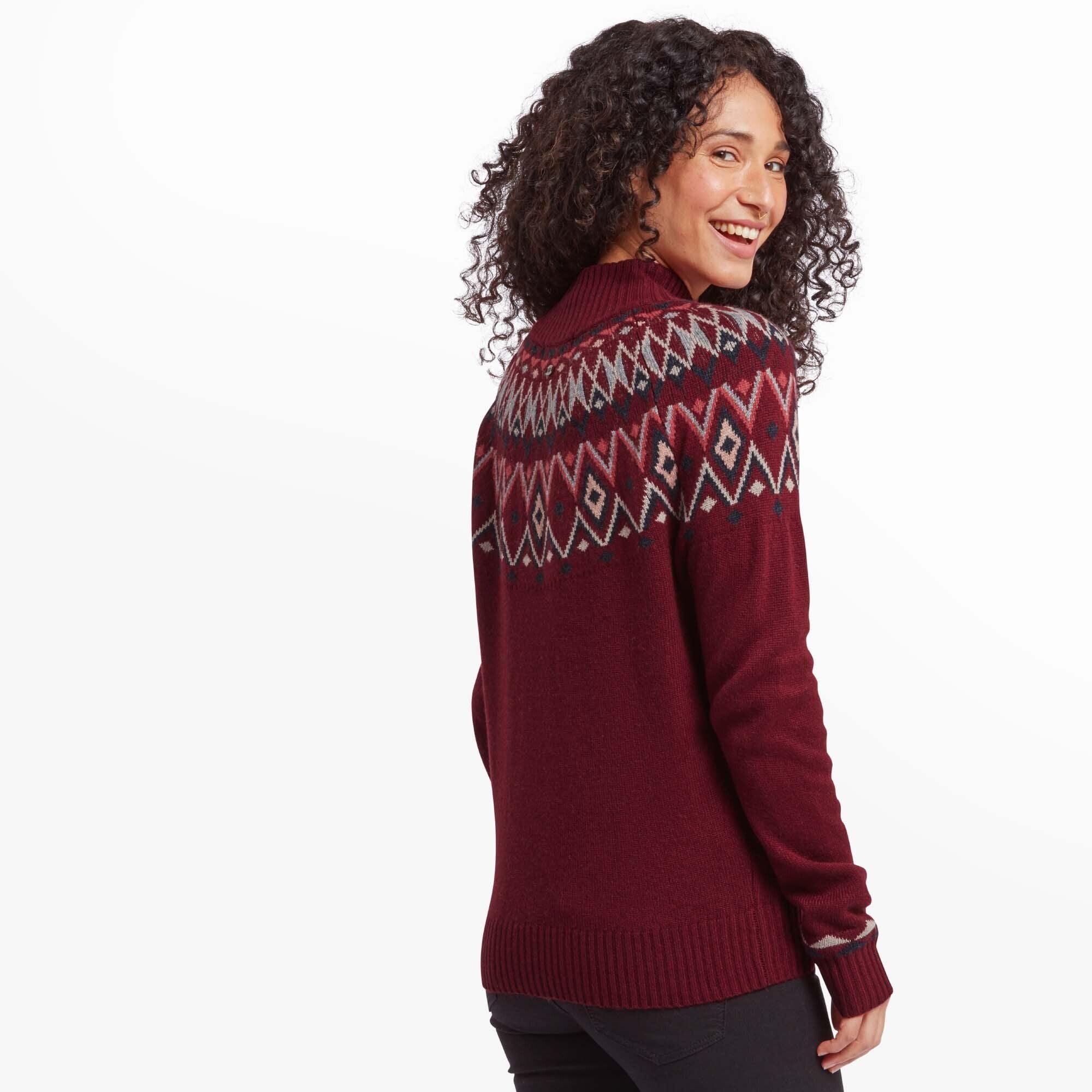 A back view of the woman in the Sherpa Adventure Gear Indu Eco Mock Neck Jumper in Red, showcasing the geometric Fair Isle design that wraps around the upper portion of the sweater. She looks over her shoulder with a cheerful expression, her curls cascading down her back.