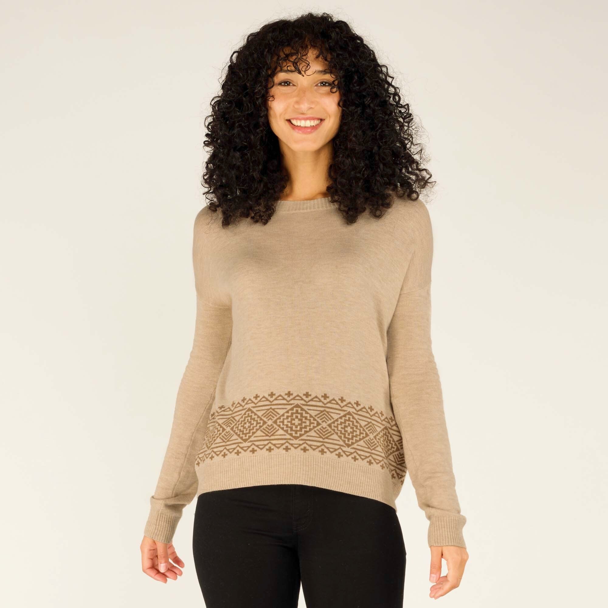 A model with curly hair smiles warmly, wearing a Sherpa Adventure Gear Jemu Crew in Beige with intricate geometric patterns in brown around the hemline. The sweater has a relaxed fit with a round neckline, paired with black fitted pants and simple black boots, standing against a neutral background.