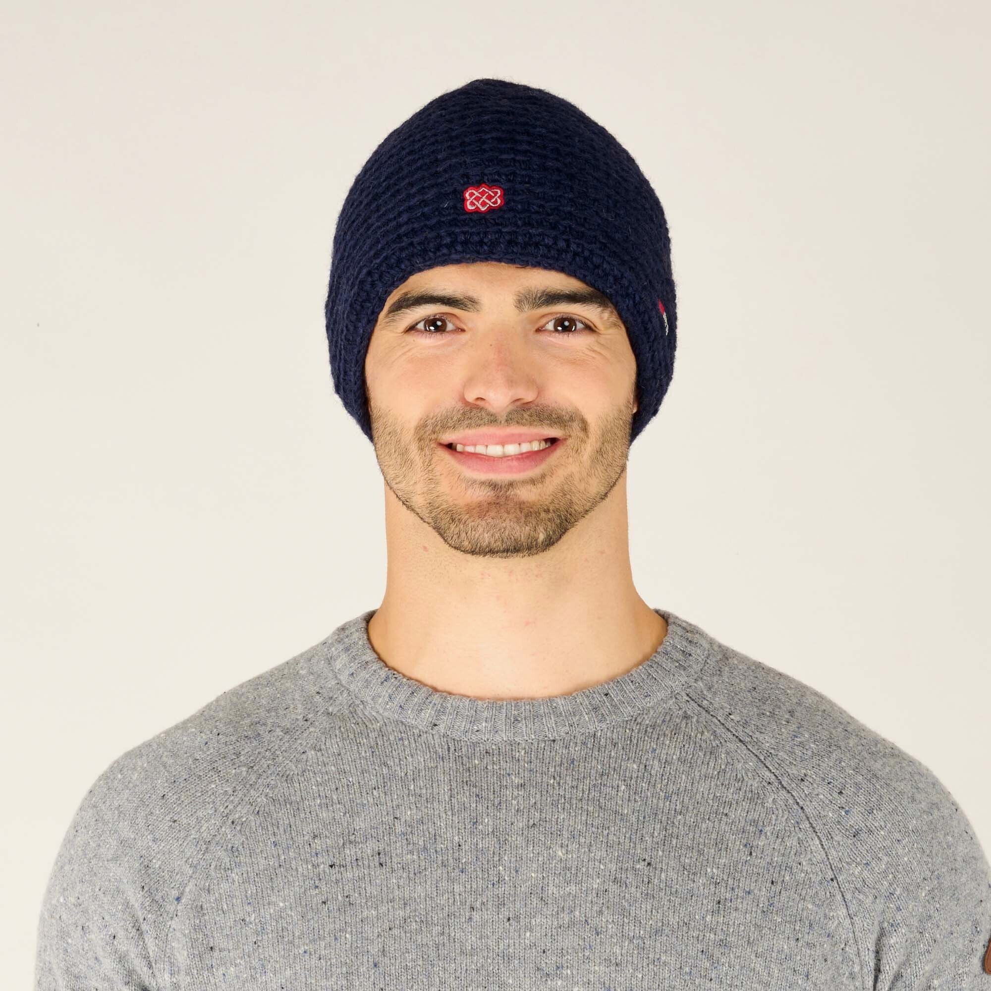 A smiling male model with light skin, short dark hair, and slight stubble wears a Sherpa Adventure Gear Jumla Hat in Blue with a red Sherpa Adventure Gear logo embroidered on the front center. The beanie has a thick, textured knit pattern. The model is also wearing a light grey speckled crew neck sweater. The background is a plain off-white studio backdrop.